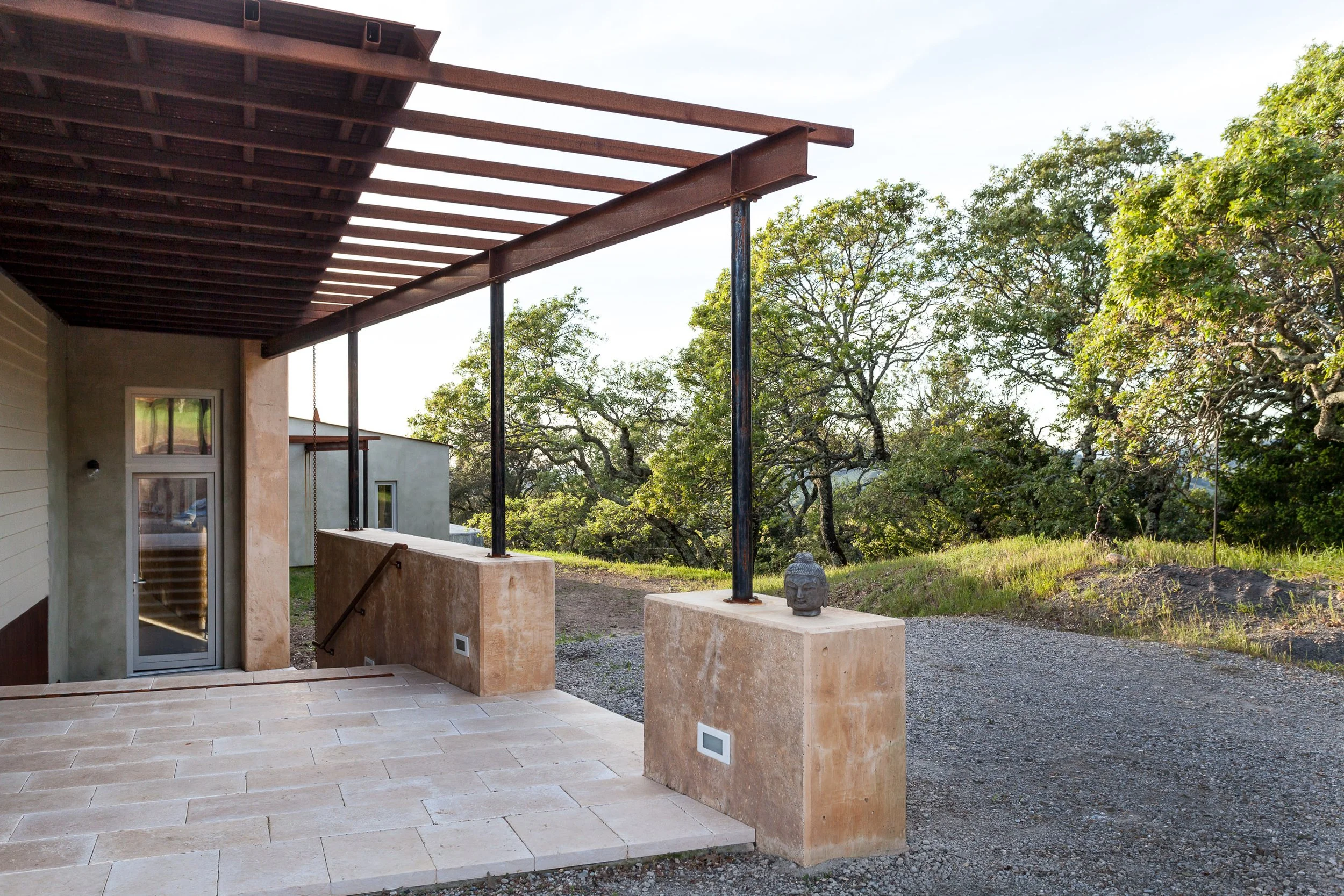 Modern patio with a pergola, stone paving, and a view of trees and grass in the background.