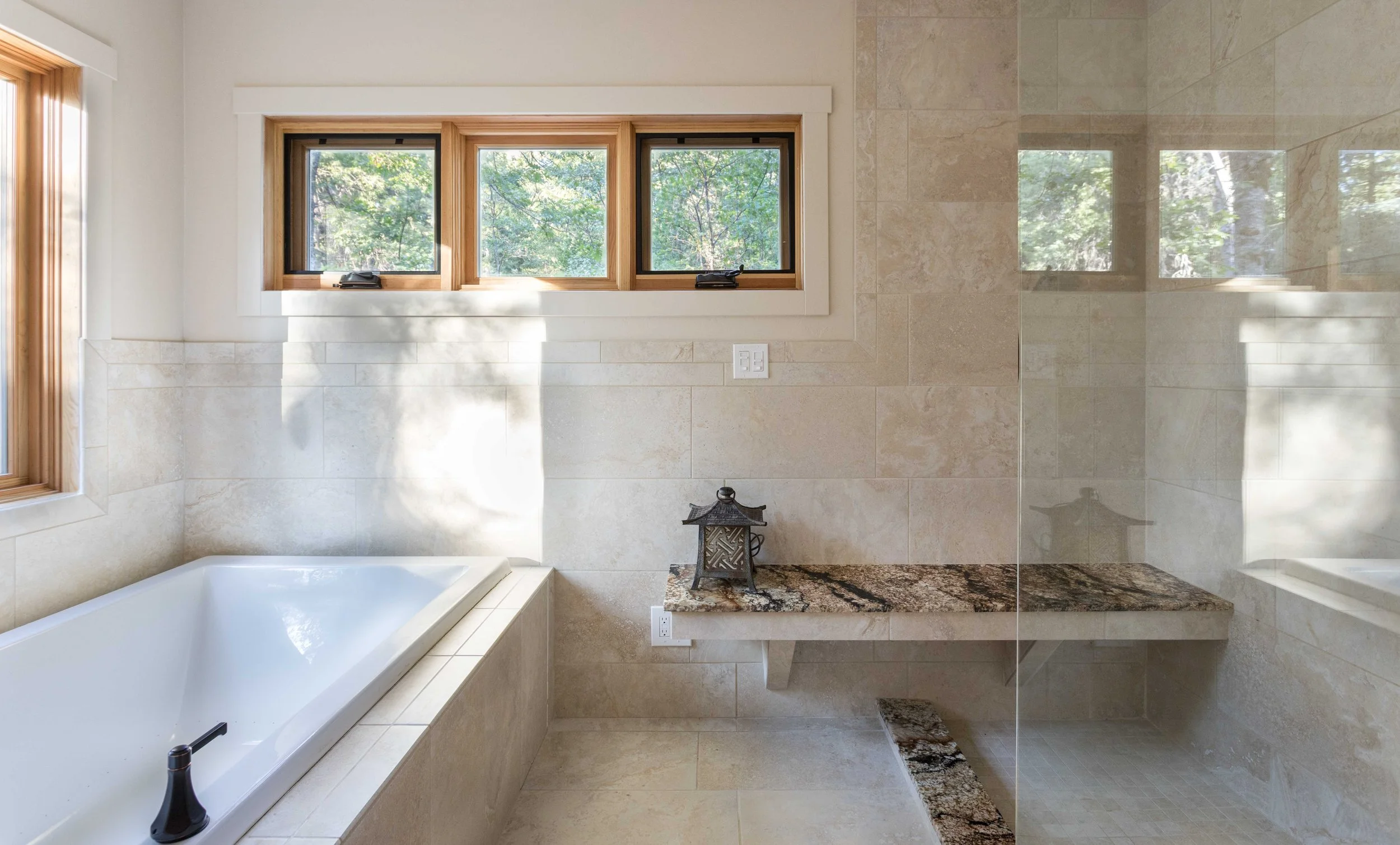 A modern bathroom with beige tiles, a white bathtub, and three small windows. There's a glass-enclosed shower area with a stone bench and a decorative lantern on a marble shelf.