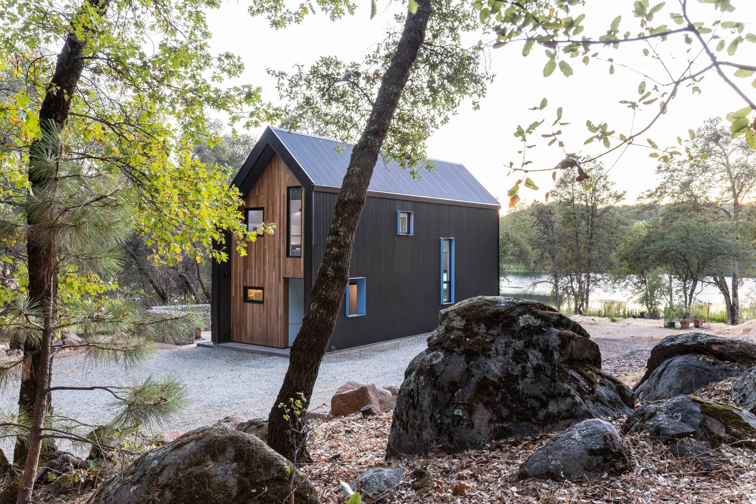 Modern cabin with black exterior, wooden accents, in a forest setting with trees, rocks, and a nearby lake.