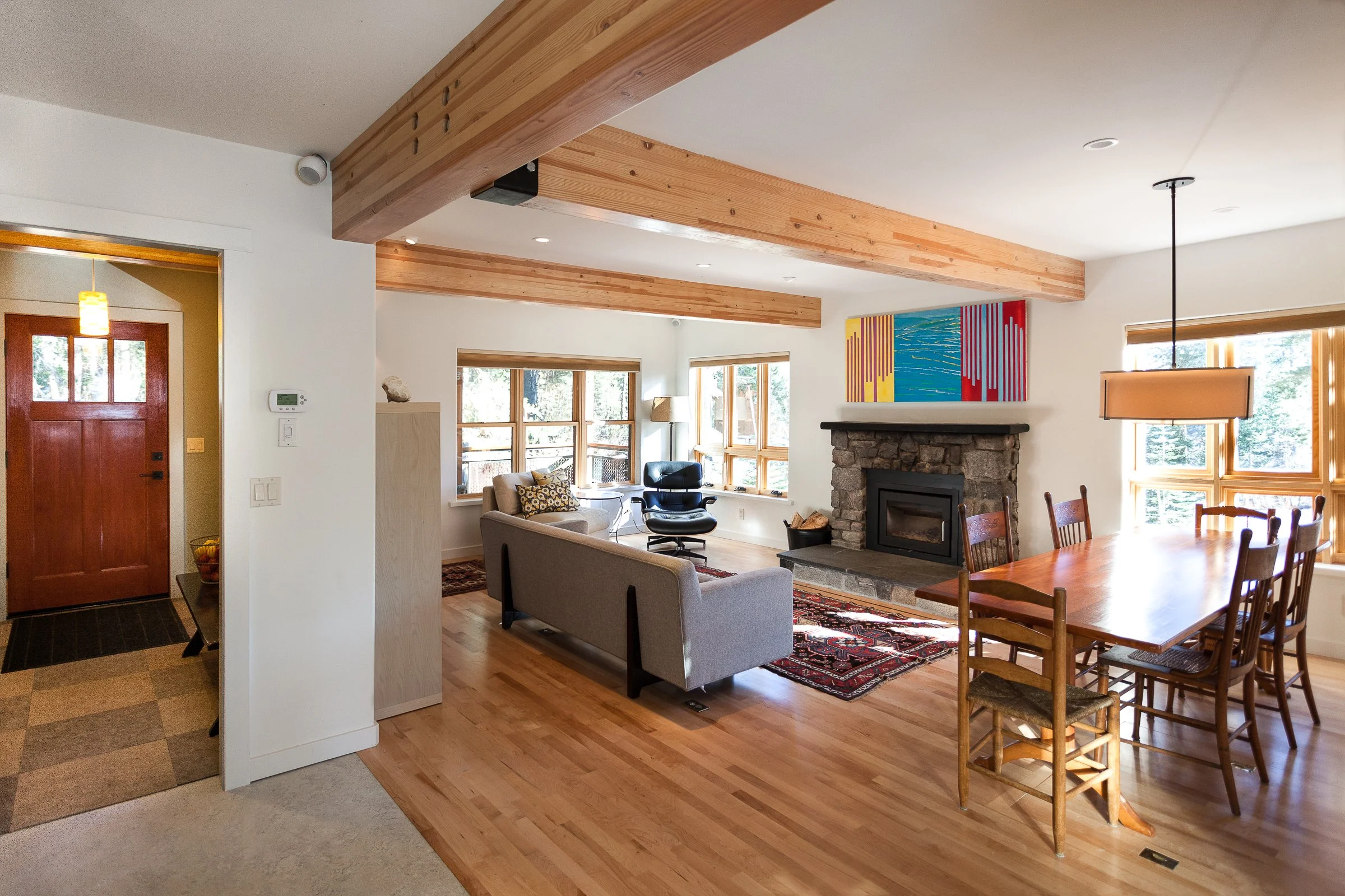 Open-concept living and dining area with wooden beams, a stone fireplace, and large windows. The room features a gray sofa, patterned rugs, wooden dining table with chairs, and colorful abstract art above the fireplace. A red front door is visible, along with light wood flooring.