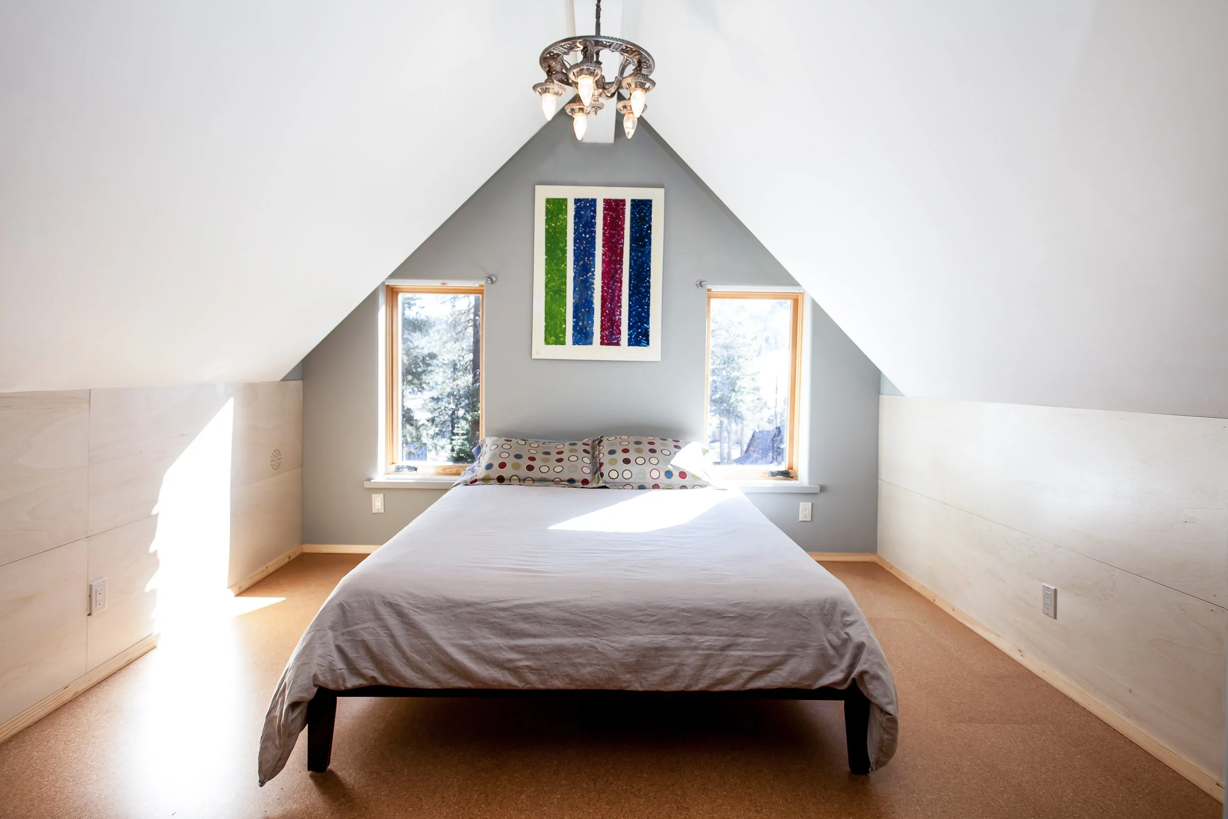 Minimalist bedroom with sloped ceiling, featuring a bed, two windows, colorful artwork, and chandelier.