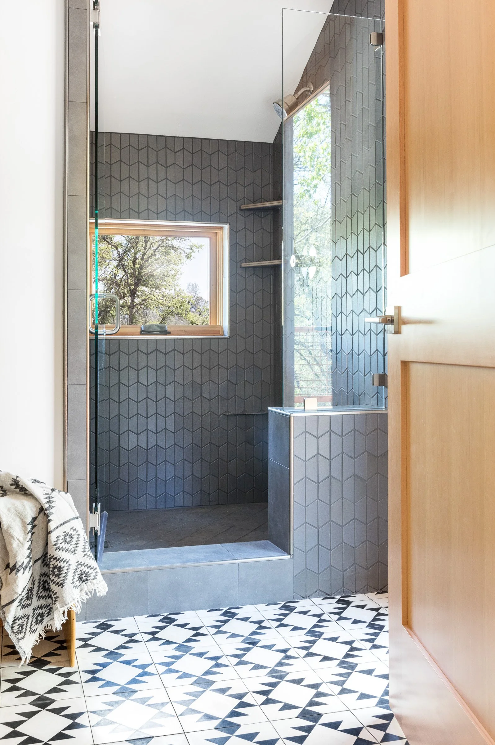Modern bathroom with geometric tiling, glass shower enclosure, wooden frame window, and patterned floor tiles.