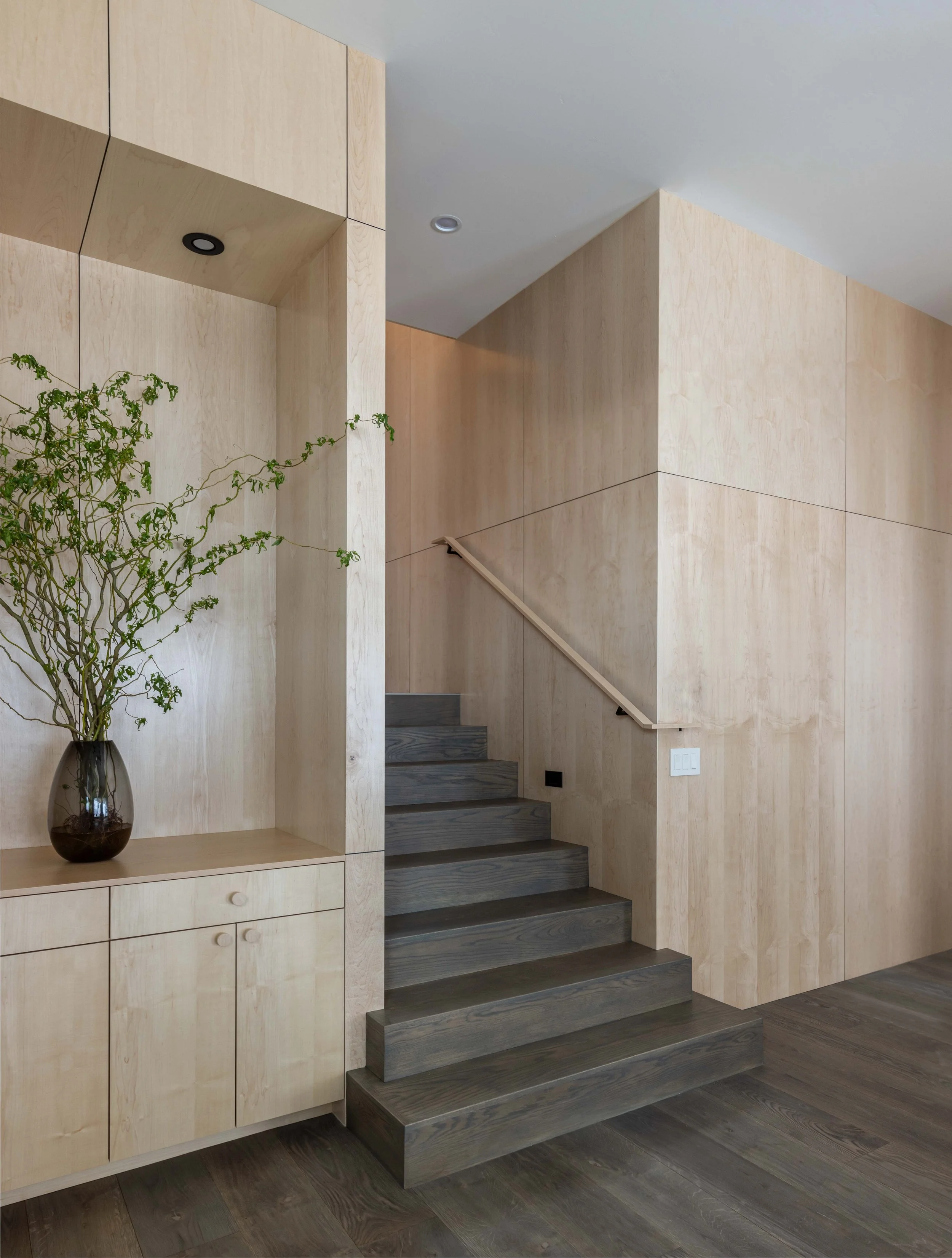 Modern minimalist interior with light wood paneling, dark wooden stairs, and a vase with branches on a shelf.