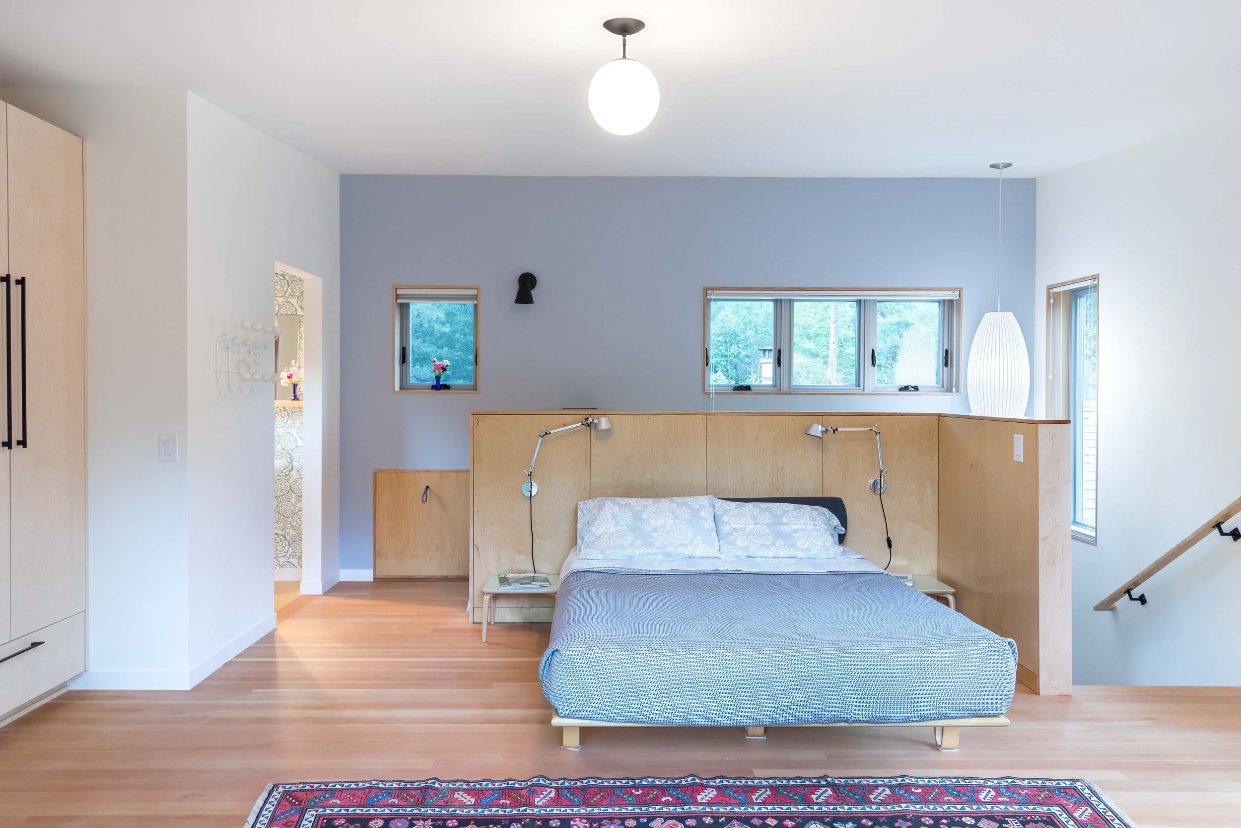 Modern bedroom with light wood flooring, a double bed with a blue-gray blanket, built-in headboard with adjustable reading lamps, and light wood paneled walls. Windows provide natural light, and a pendant light hangs above. A colorful rug is on the floor, and there are doors leading to other areas.