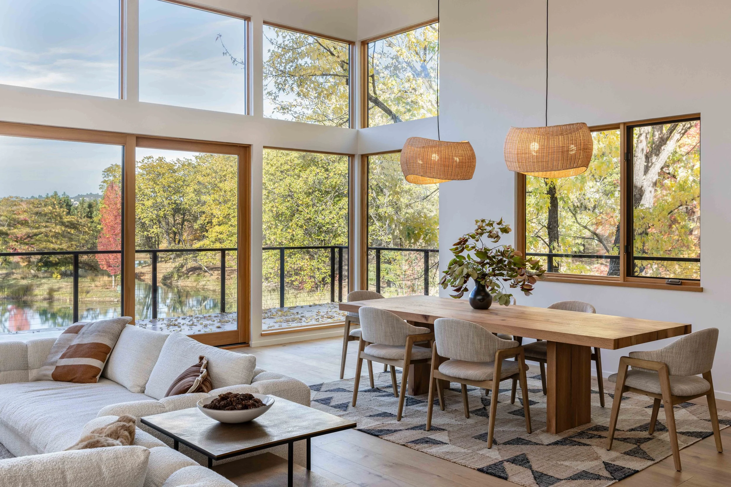 Modern living and dining room with large windows overlooking a forest view, featuring a white sectional sofa, wooden dining table, beige chairs, patterned rug, and woven pendant lights.