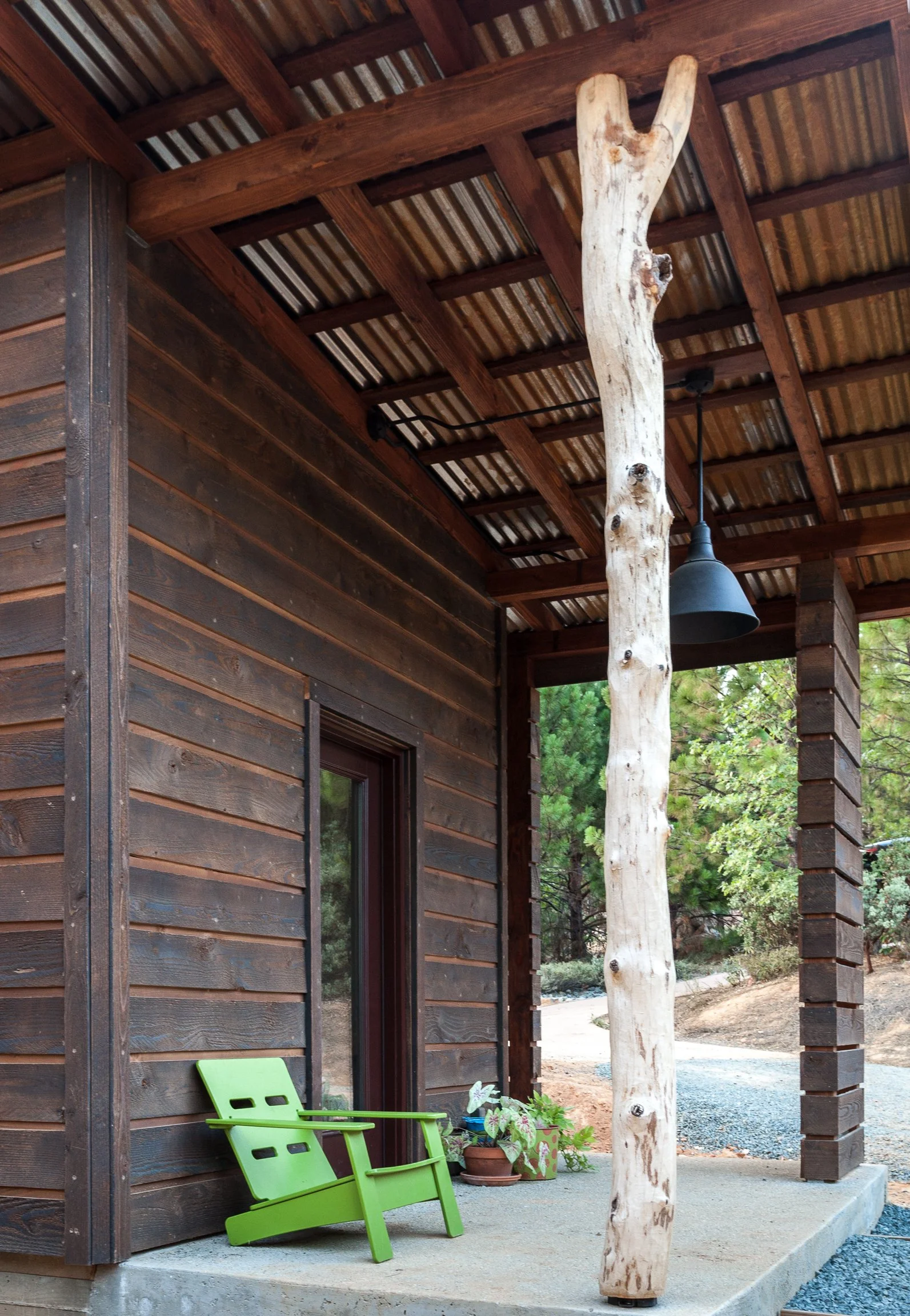 Rustic wooden porch with corrugated metal roof, natural log support post, green Adirondack chair, potted plants, and surrounding greenery.