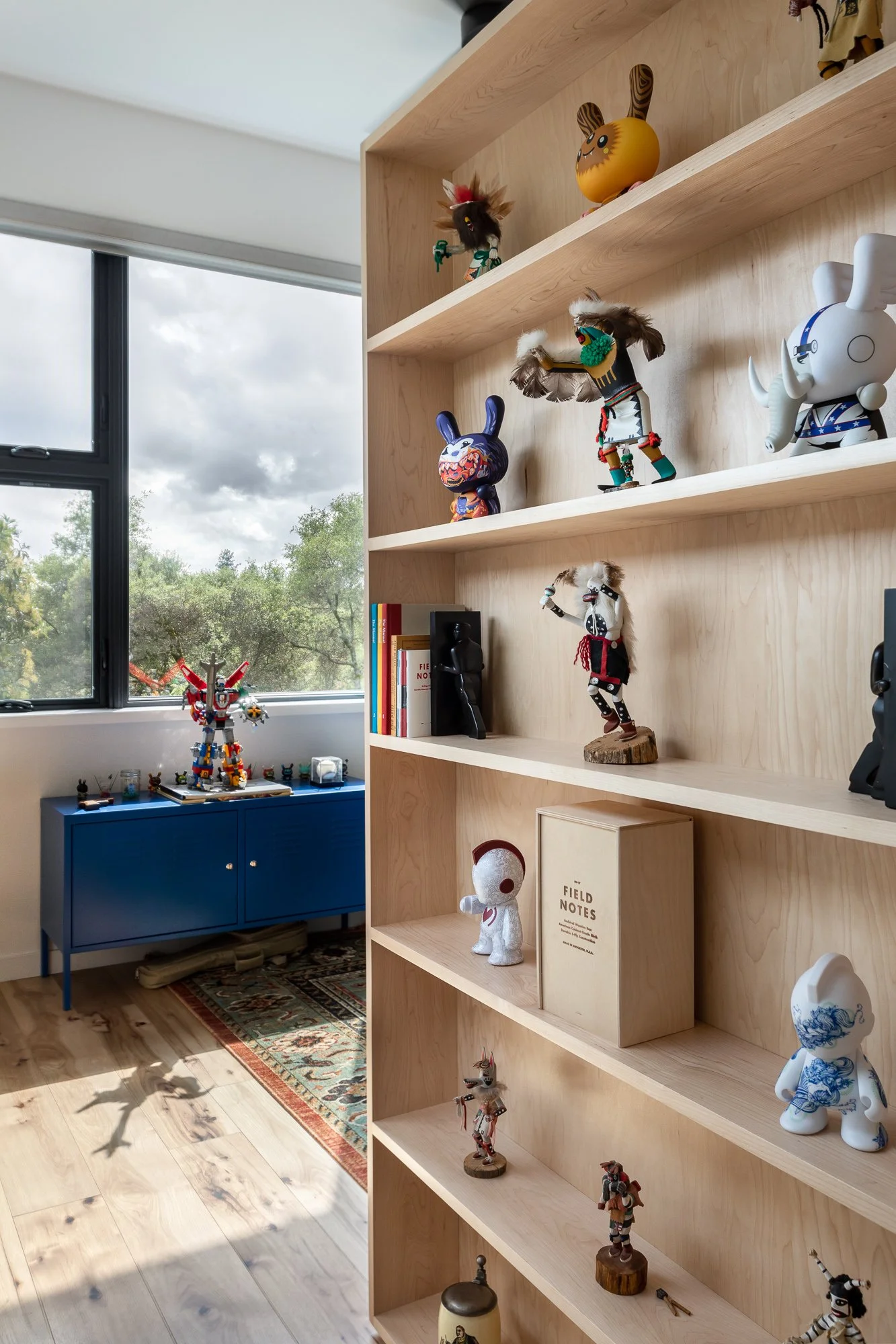 Room with a wooden bookshelf displaying various figurines and toys, blue cabinet by the window, natural light coming in, and a decorative rug on the wooden floor.