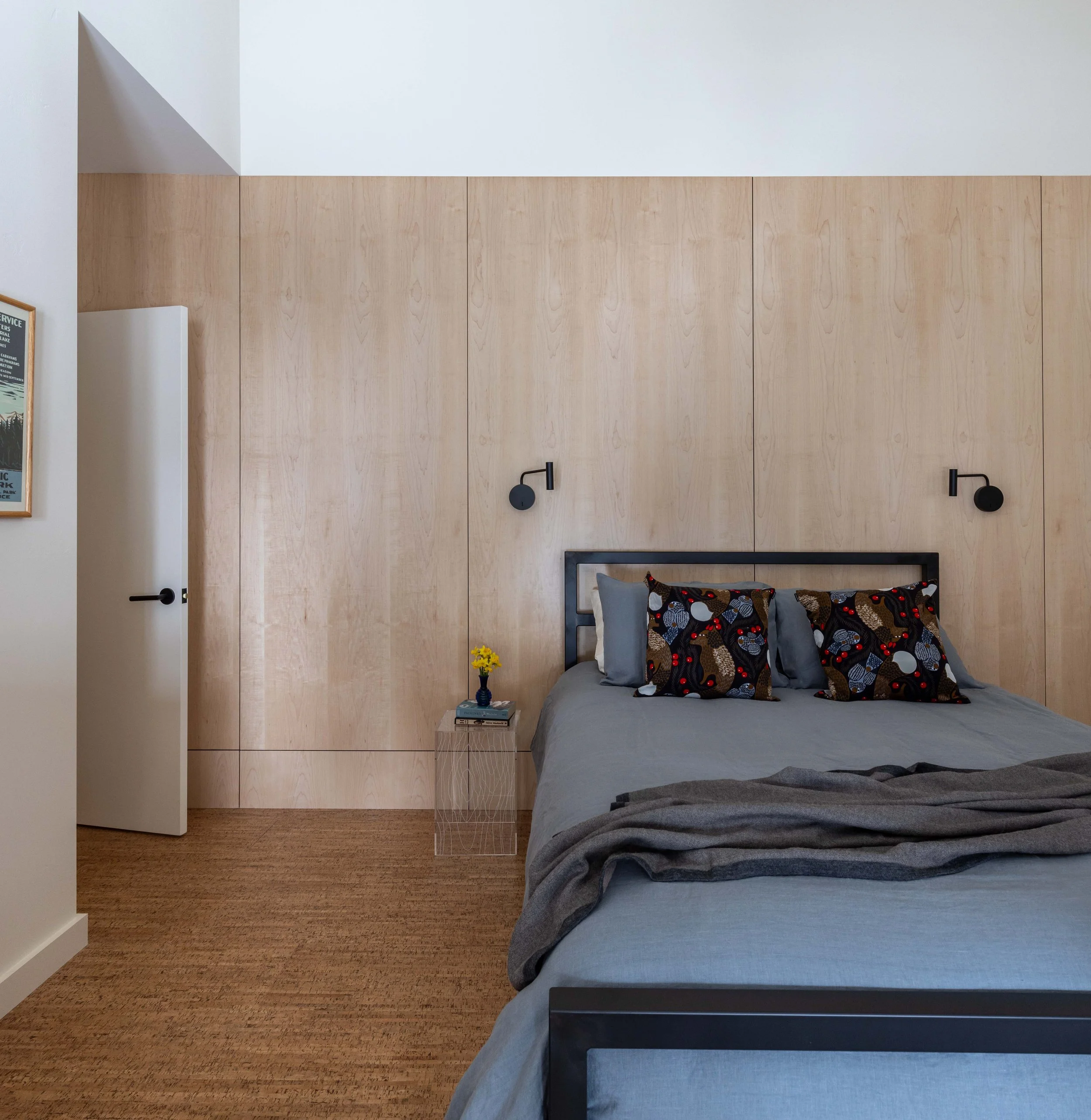 Modern bedroom with wood panel wall, gray bedding, decorative pillows, wall-mounted reading lights, and a side table with a flower vase.