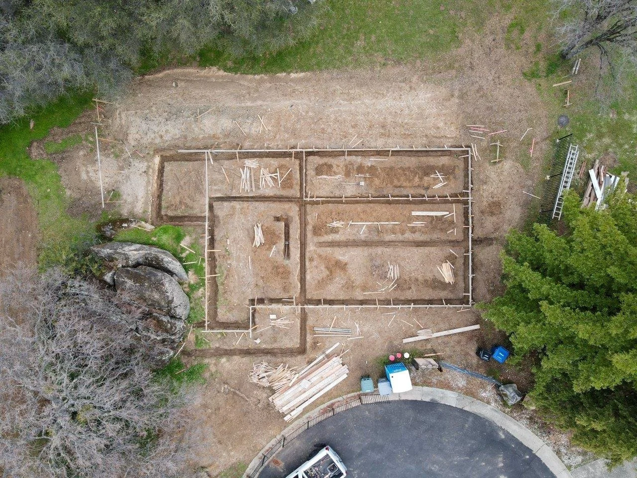 Aerial view of a construction site with a foundation layout, surrounded by trees and rocks, and adjacent to a paved road.