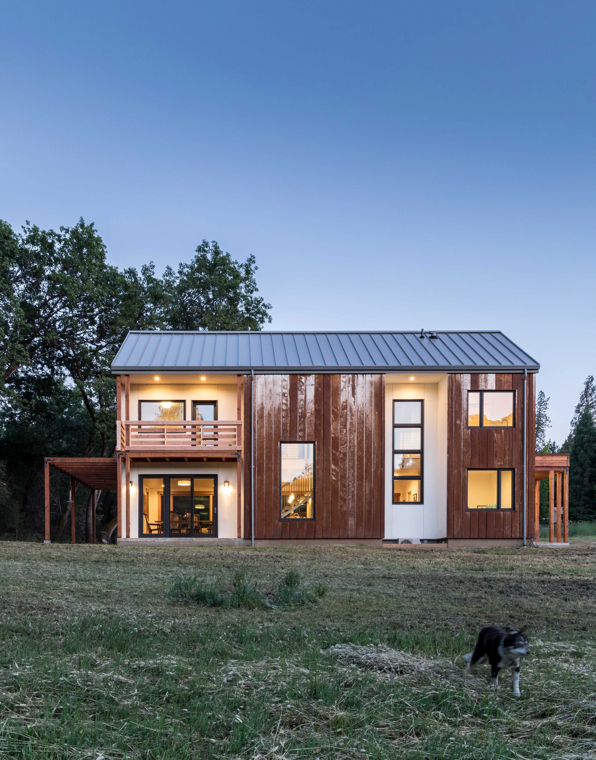Modern two-story wooden house with large windows, a balcony, and a metal roof. The house is surrounded by trees and open grassland, with a dog playing in the foreground during dusk.