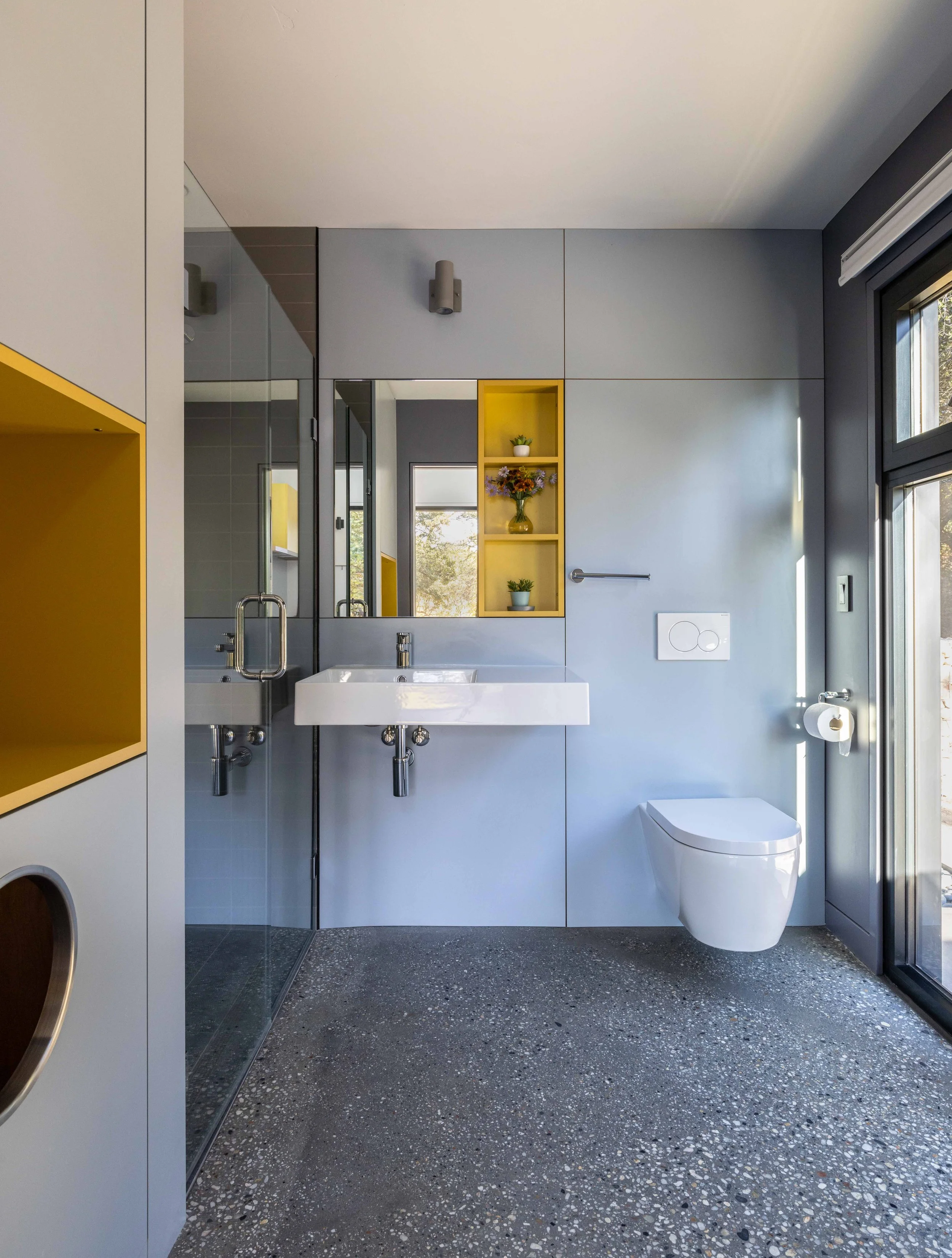 Modern bathroom with minimalist design, featuring a wall-mounted toilet, rectangular sink, large mirror, glass shower enclosure, and yellow shelving for decor. The room has gray walls and concrete flooring with a window for natural light.