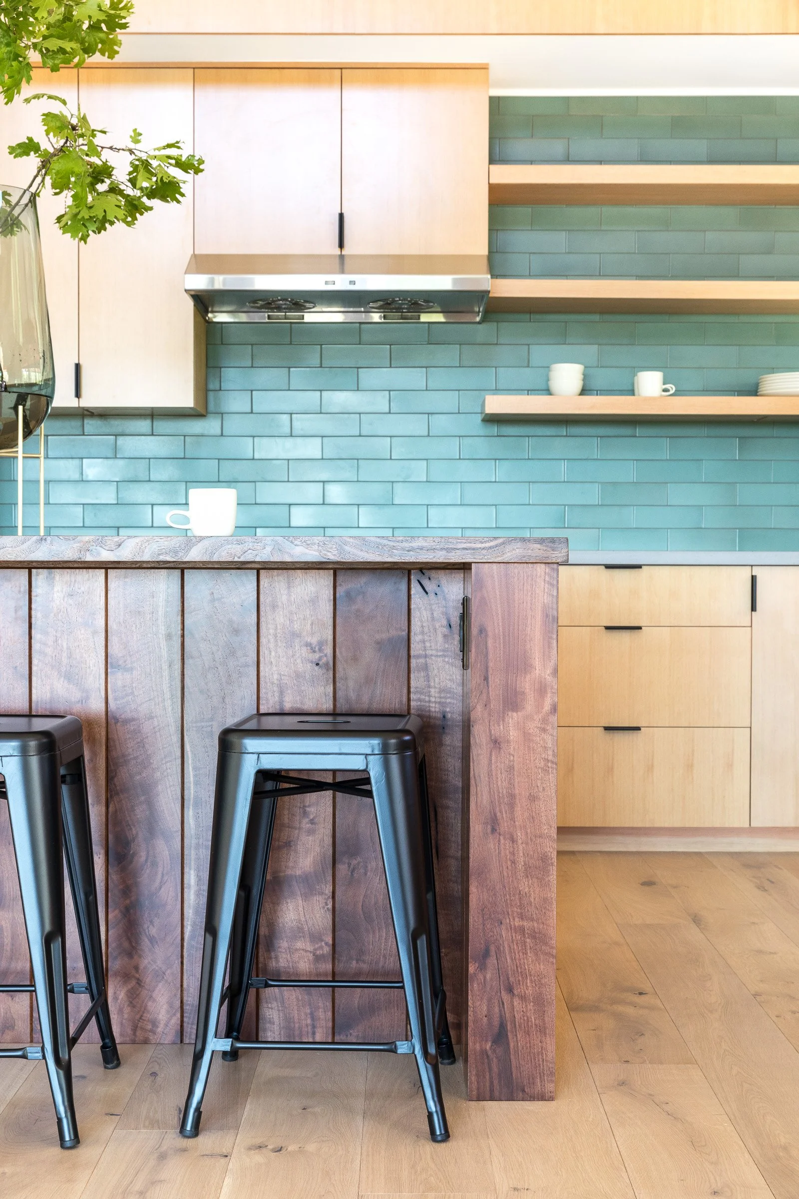 Modern kitchen with teal subway tile backsplash, light wood cabinetry, open shelves with white cups, and a wood-paneled island with black metal stools.