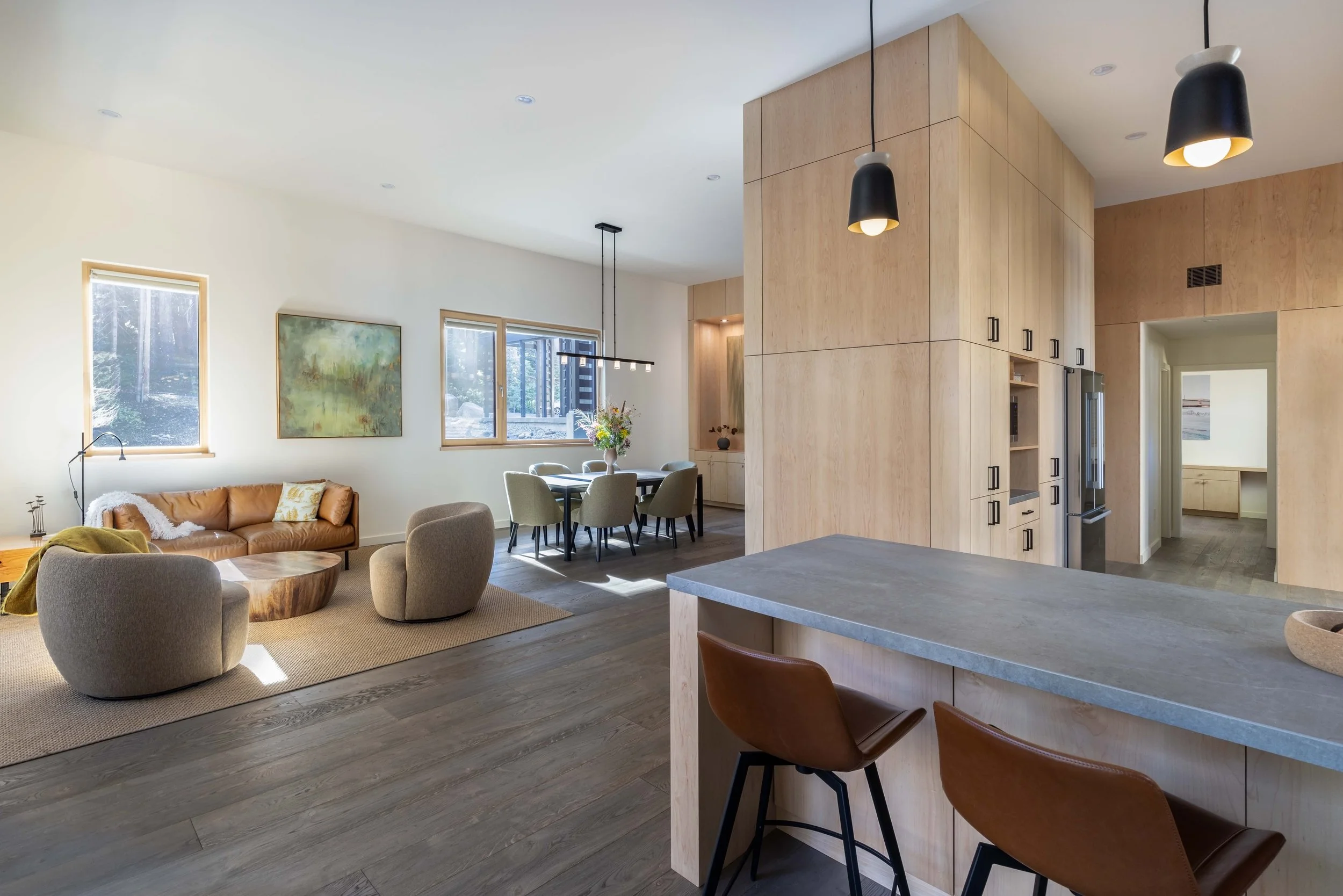Modern open-concept living space with a beige sofa, armchairs, and a wooden coffee table in the living area. A dining table with chairs and a large wooden cabinet are visible in the middle. Pendant lights hang above a kitchen island with leather stools.