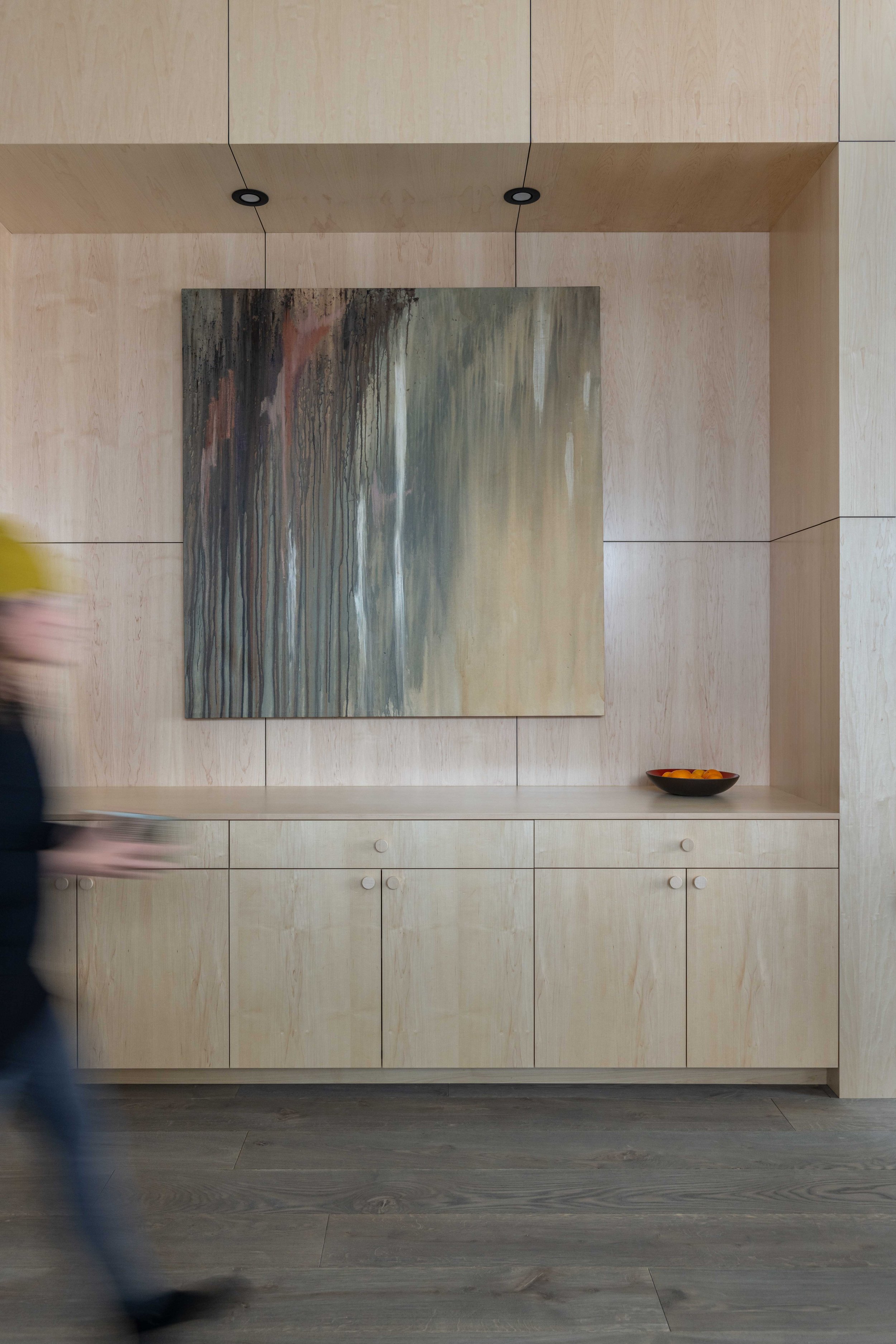 Blurred person walking past a modern wooden cabinet with an abstract painting and a bowl of fruit on top.