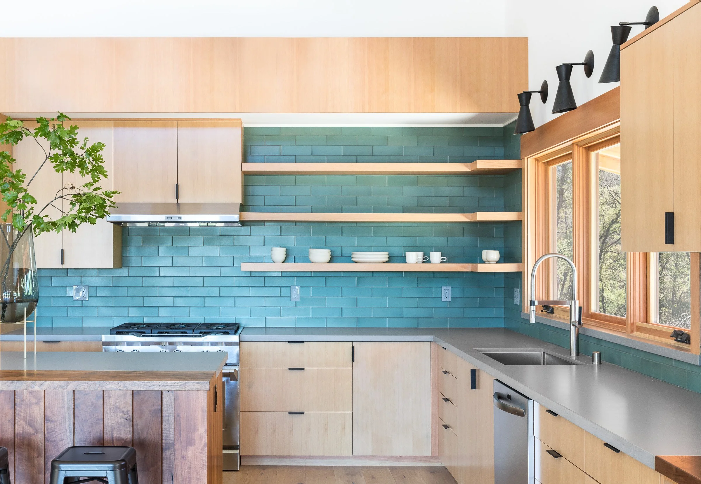 Modern kitchen with light wood cabinets, teal subway tile backsplash, open shelves with white dishes, stainless steel appliances, and a large window.
