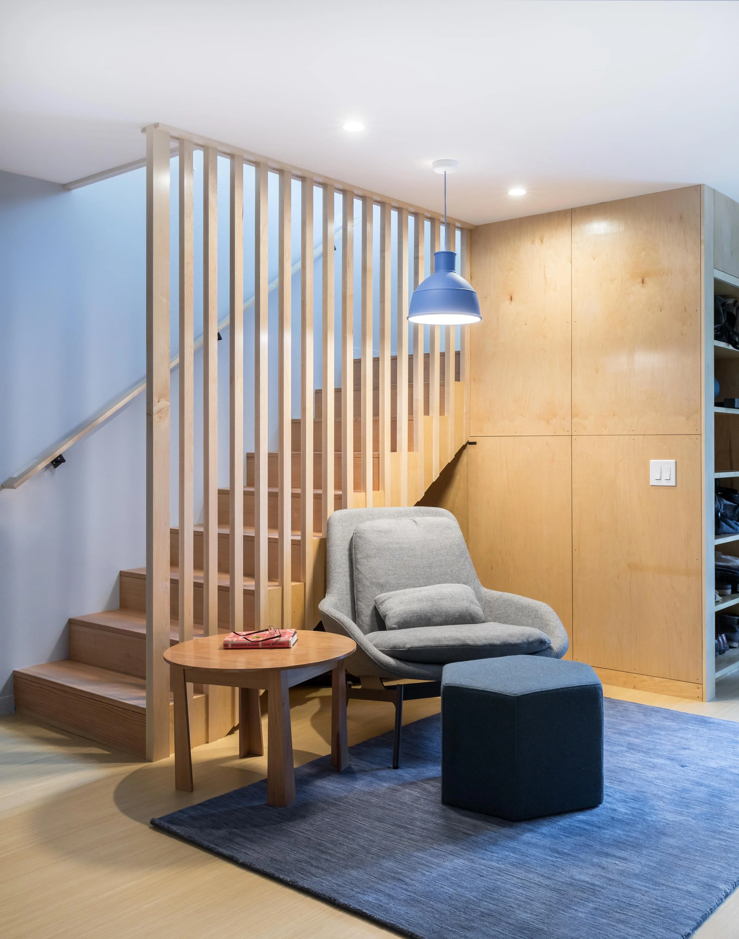 Modern interior with a gray armchair, wooden table, and blue pouf on a blue rug, under a blue pendant light, next to wooden stairs with vertical slats.