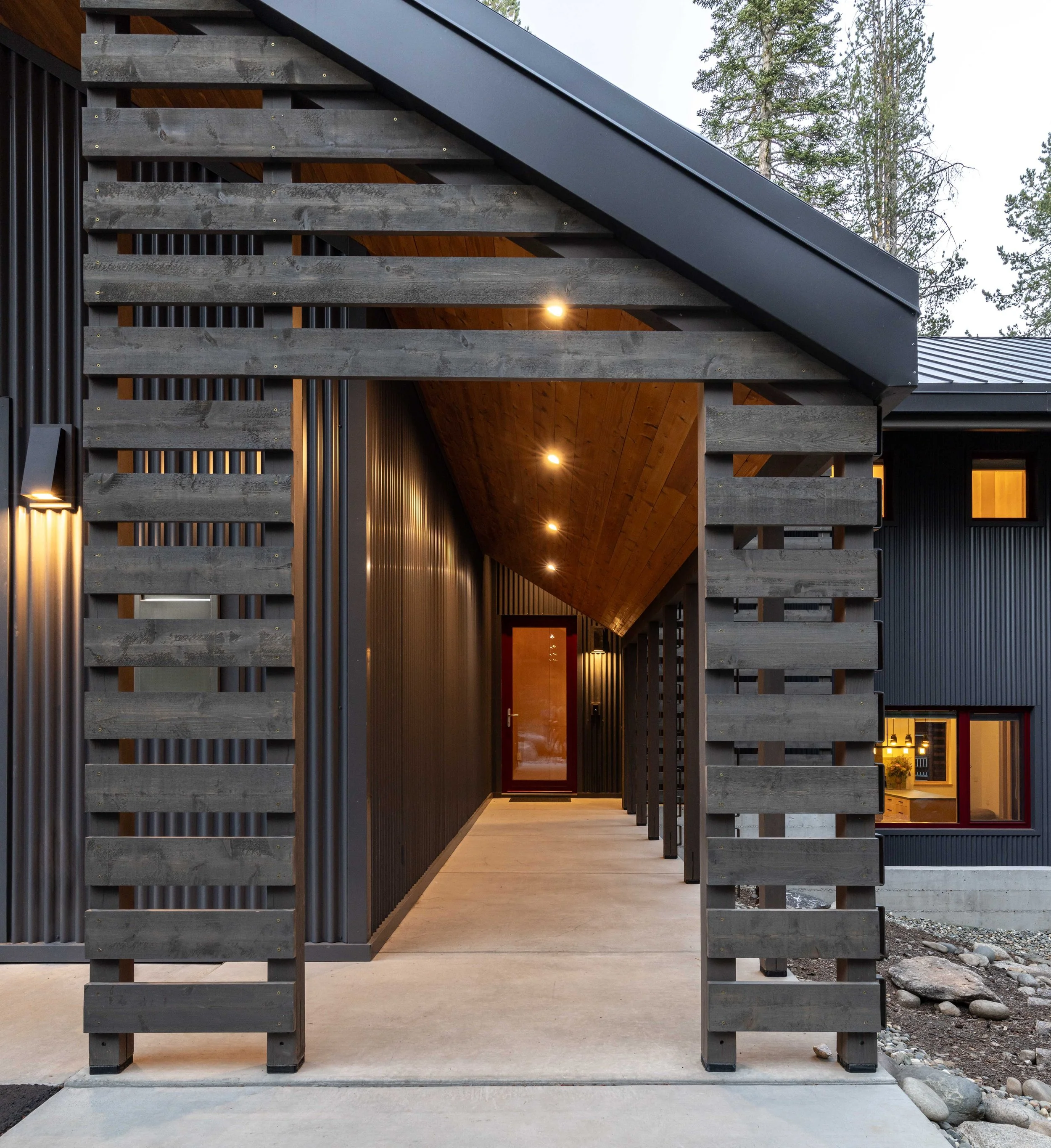 Contemporary house entrance with a wooden pergola and corrugated metal siding, featuring illuminated ceiling lights and a glass door at the end of the walkway.