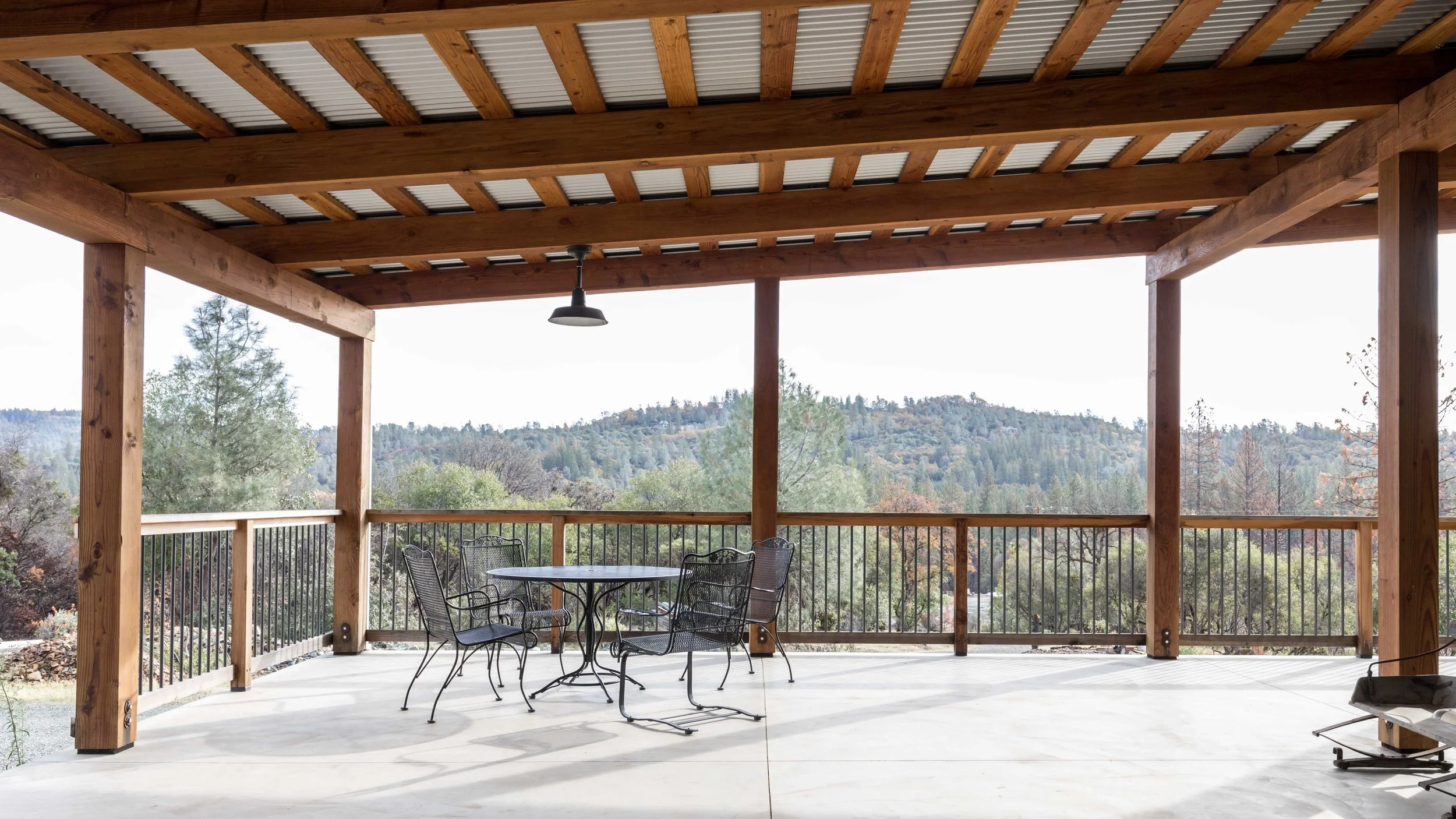 Outdoor covered patio with wooden beams, metal railing, round table, and chairs, overlooking a forested mountain landscape.