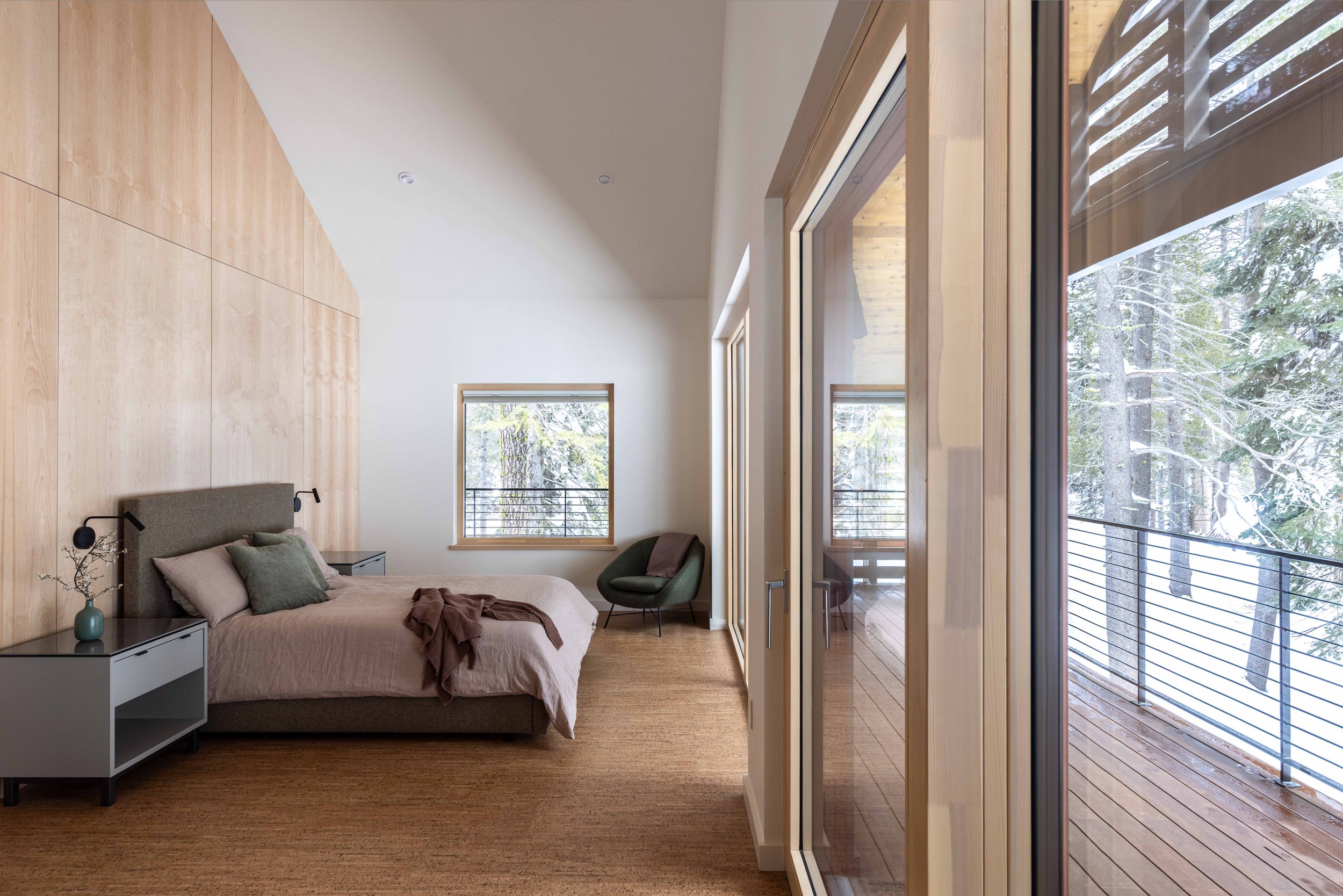 Modern bedroom with large window and wooden elements, featuring a bed with gray and green pillows, a small nightstand, a cushioned chair, and access to an outdoor patio.