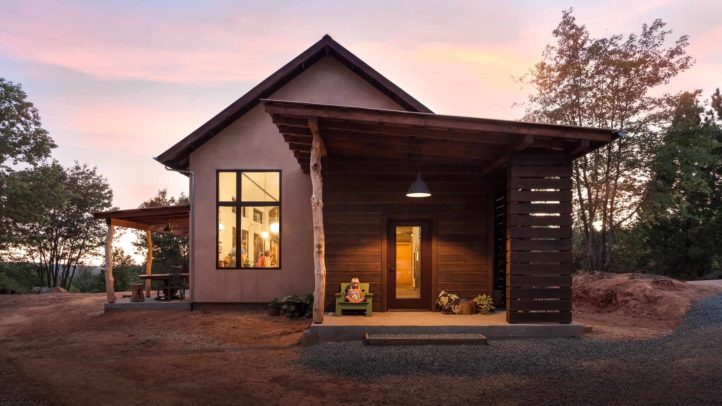Small rustic house at sunset with wide windows and natural wooden beams, surrounded by trees and gravel path.