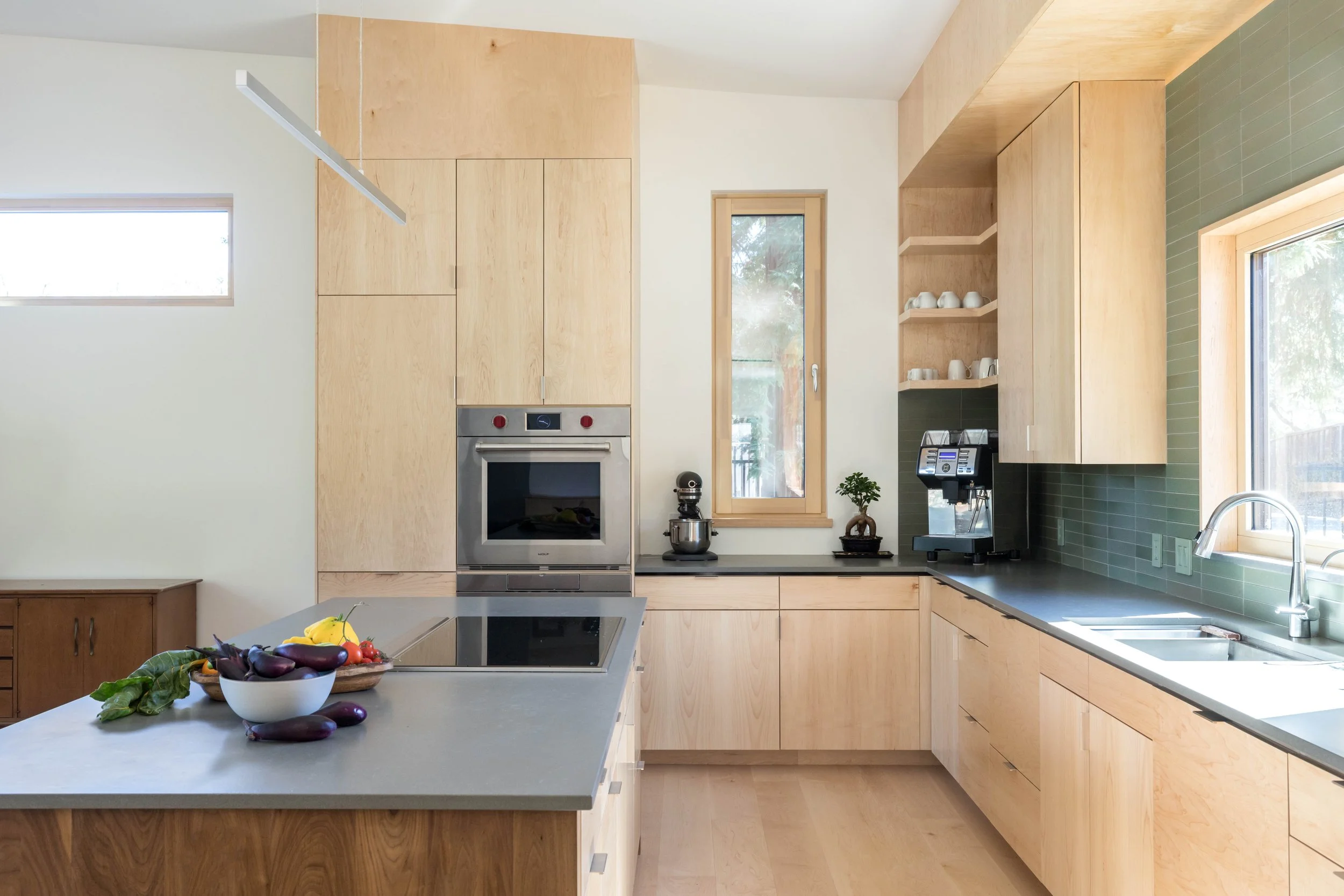 Modern kitchen with light wood cabinets, stainless steel appliances, green tile backsplash, island countertop with fresh vegetables, and small potted plant.