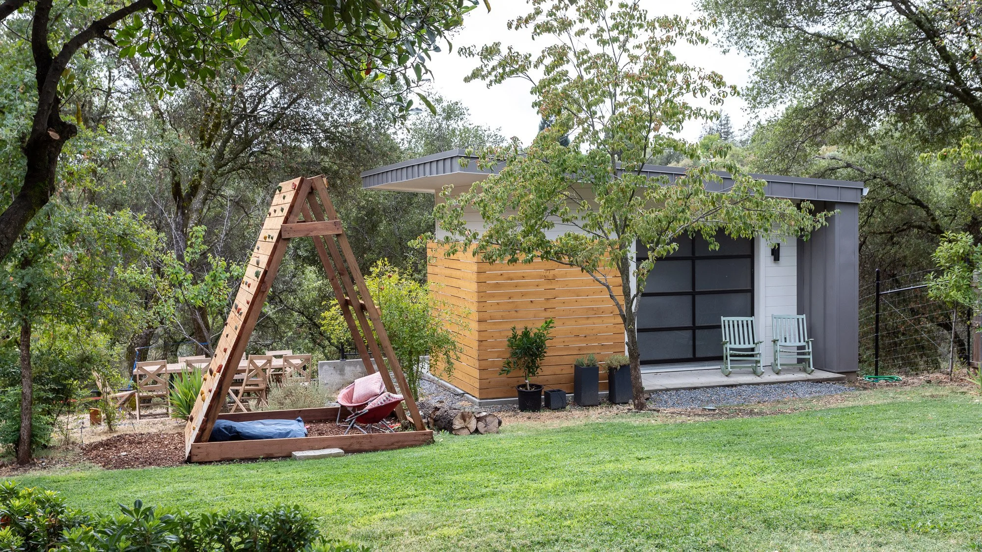 Outdoor scene with a small modern wooden cabin with large windows, two green chairs on the porch, a wooden A-frame structure for climbing, green grass, and various trees and plants surrounding the area.