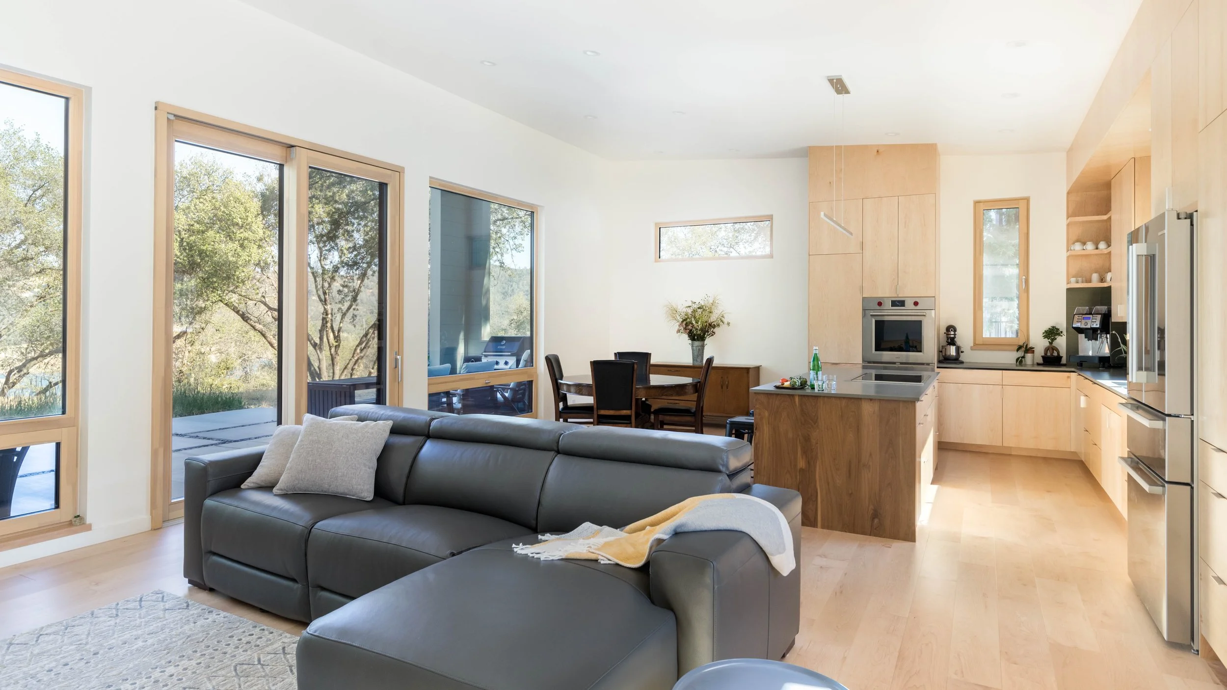 Modern open-concept living room and kitchen with large windows, gray sectional sofa, wooden dining table, and light wood cabinetry.