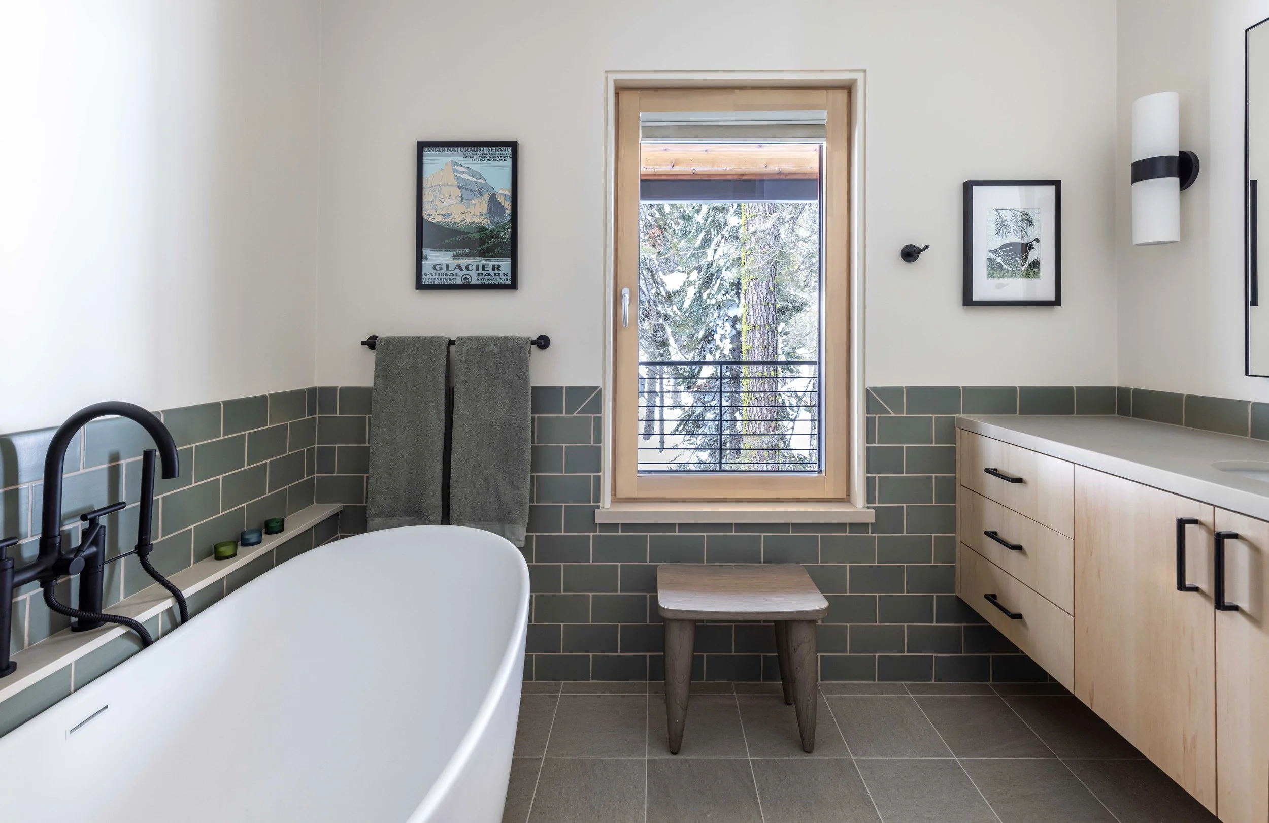 Modern bathroom with freestanding bathtub, light wood vanity, green tiled walls, and window view of snowy trees.