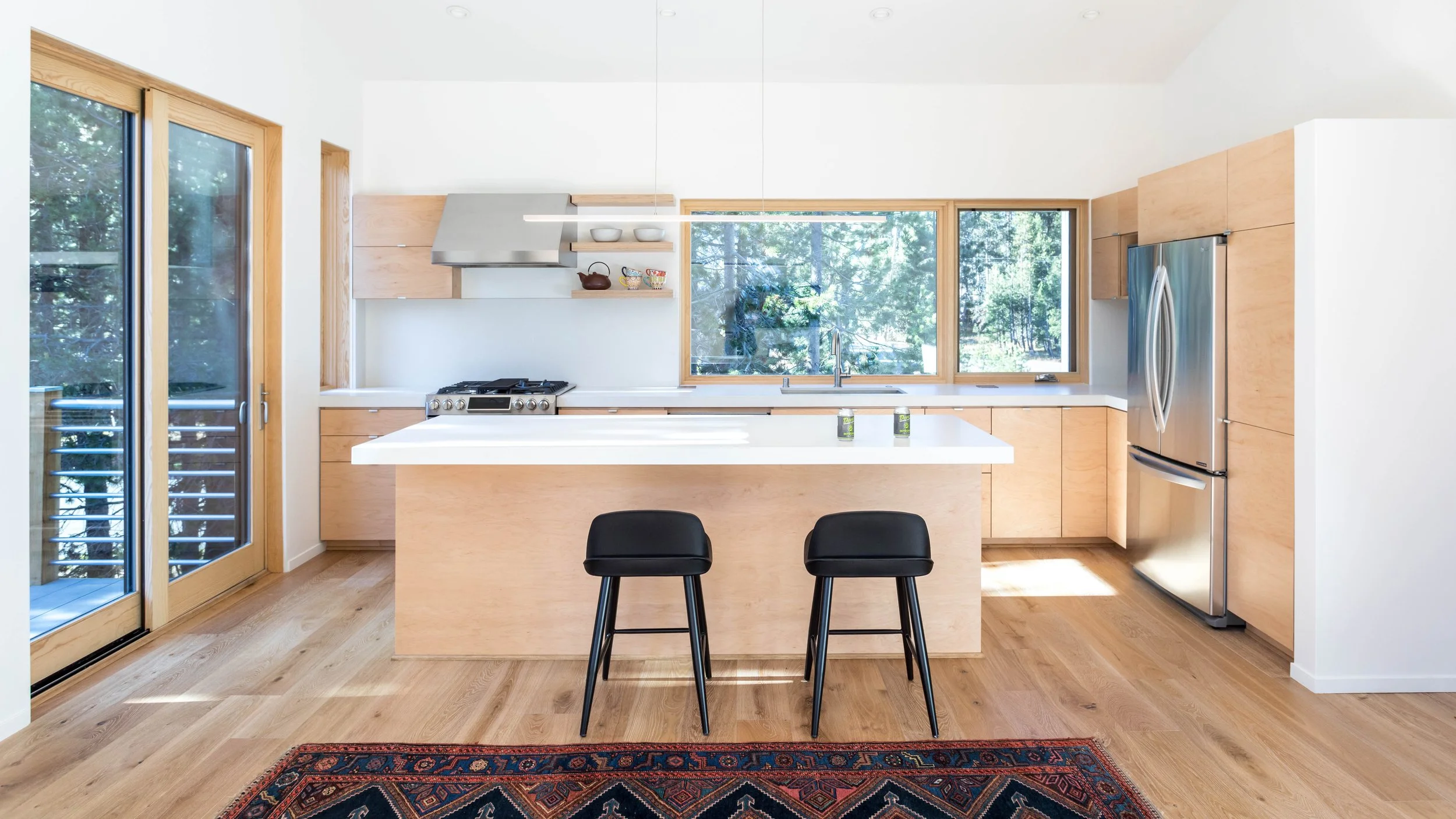 Modern kitchen with light wood cabinets, an island with two black stools, stainless steel appliances, large window, and sliding glass doors.