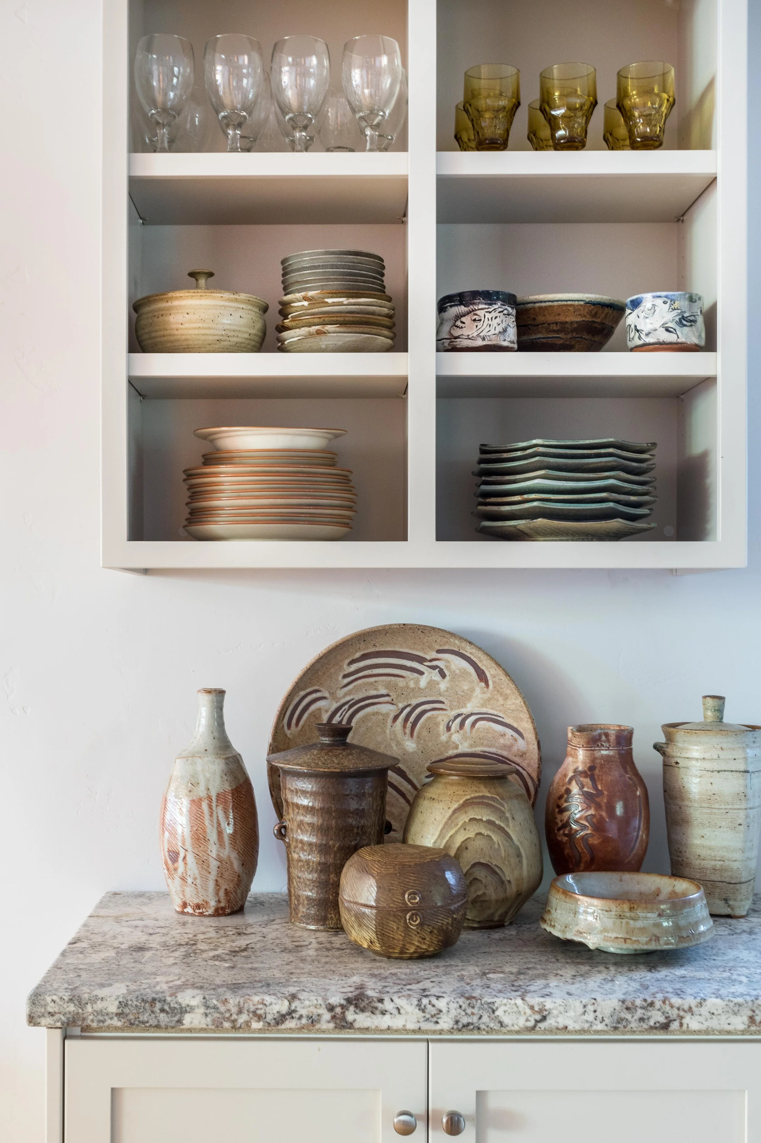 Kitchen shelves with assorted ceramic dishes, plates, bowls, and glassware. Countertop with various pottery and a large decorative plate in earth tones.