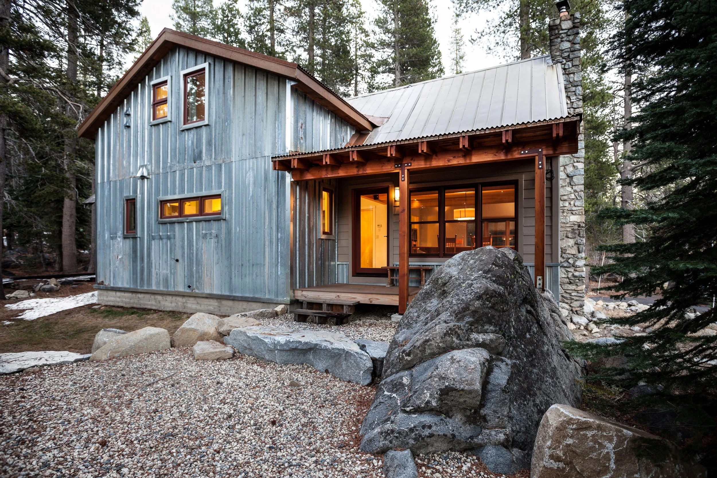 Rustic cabin with wooden and metal siding, glowing interior lights, porch, and stone chimney, surrounded by trees and rocks.