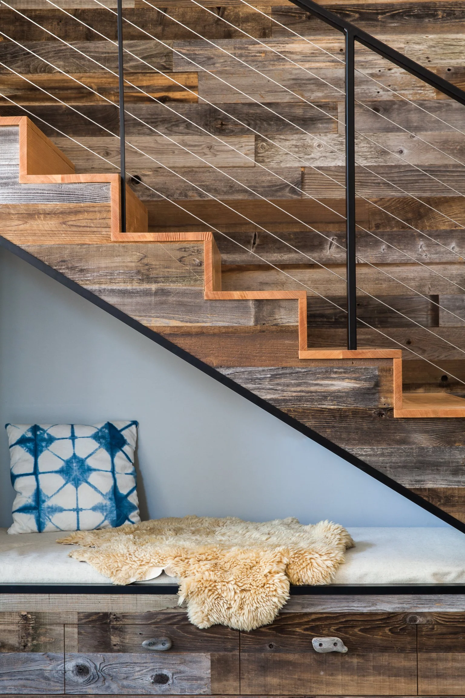 Wooden staircase with wire railing above a cozy nook with a blue patterned pillow and a fluffy beige throw.