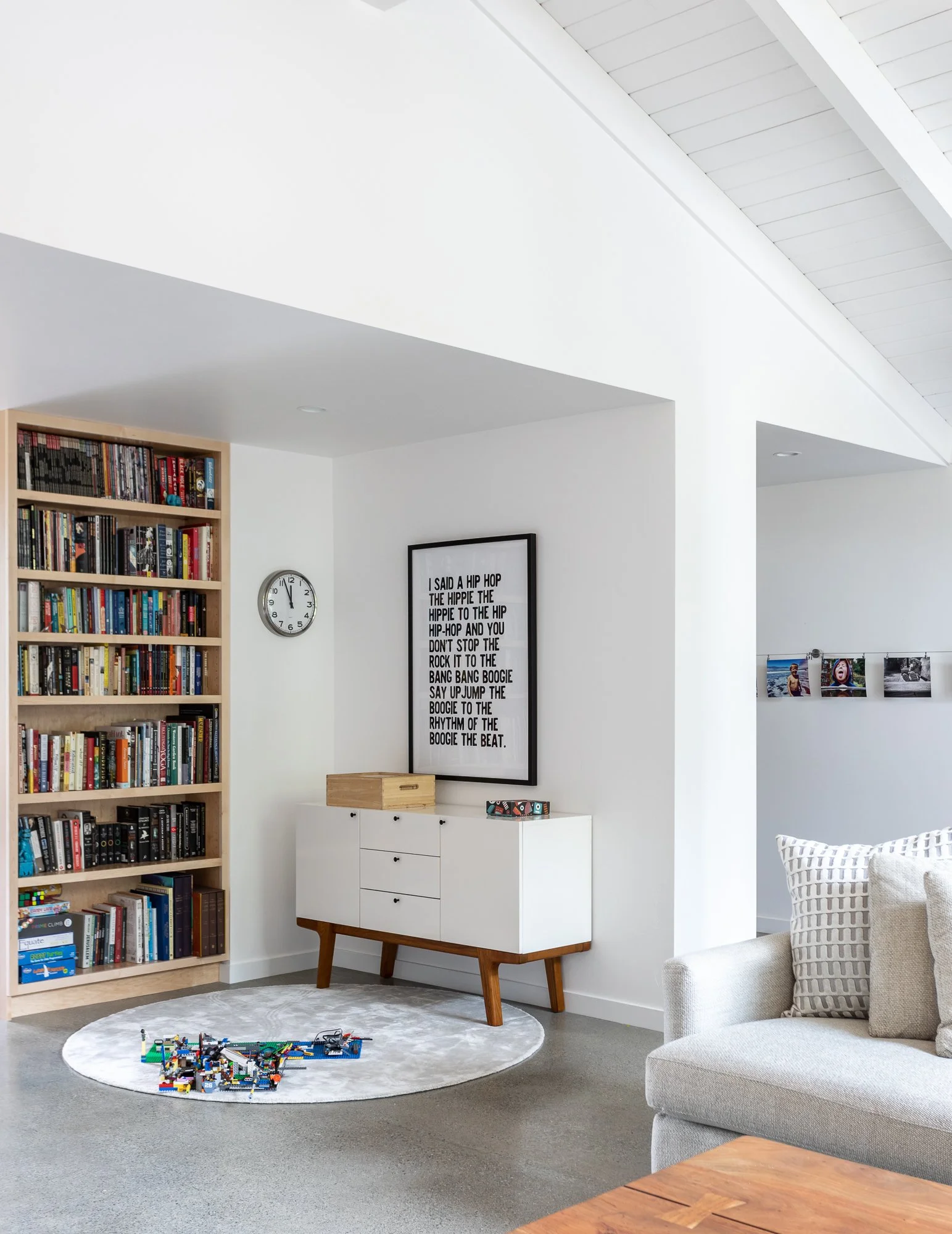 Modern living room with a bookshelf filled with books, a wall clock, a framed artwork with song lyrics, a white sideboard, a small round rug with toys, and a gray couch with patterned cushions.