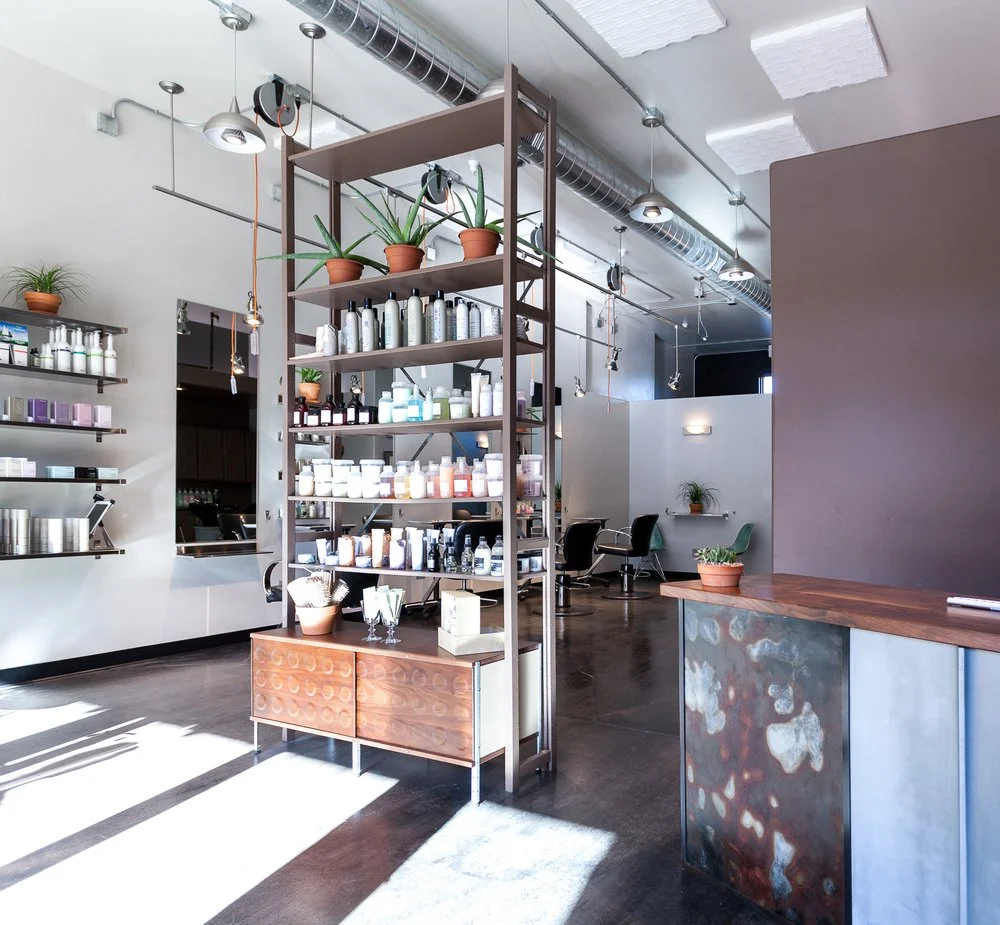 Interior of a modern hair salon with shelves of hair products, potted plants, and styling stations with chairs.