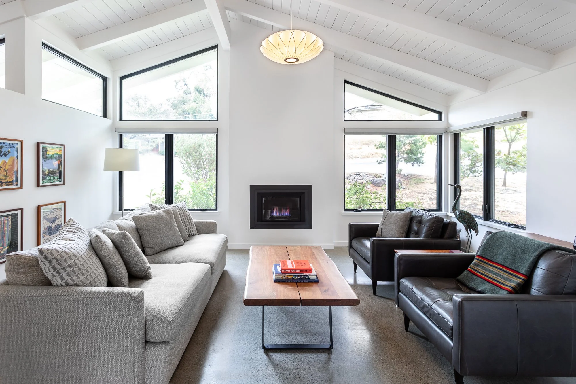 Modern living room with large gray sofa, two brown leather armchairs, wooden coffee table, wall-mounted fireplace, large windows, and decorative artwork on the walls.