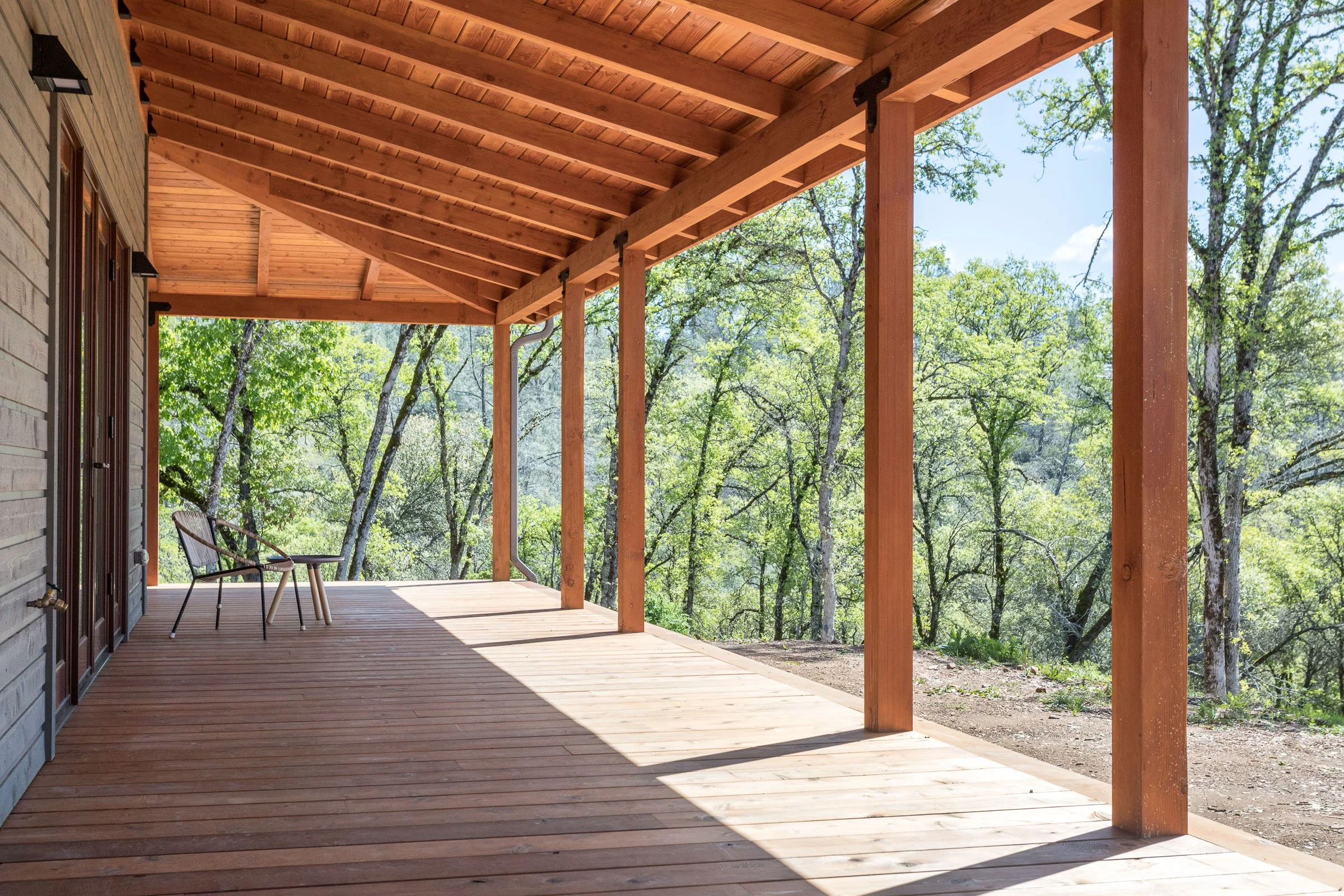 Wooden porch with roof, chair, and small table overlooking lush green trees.