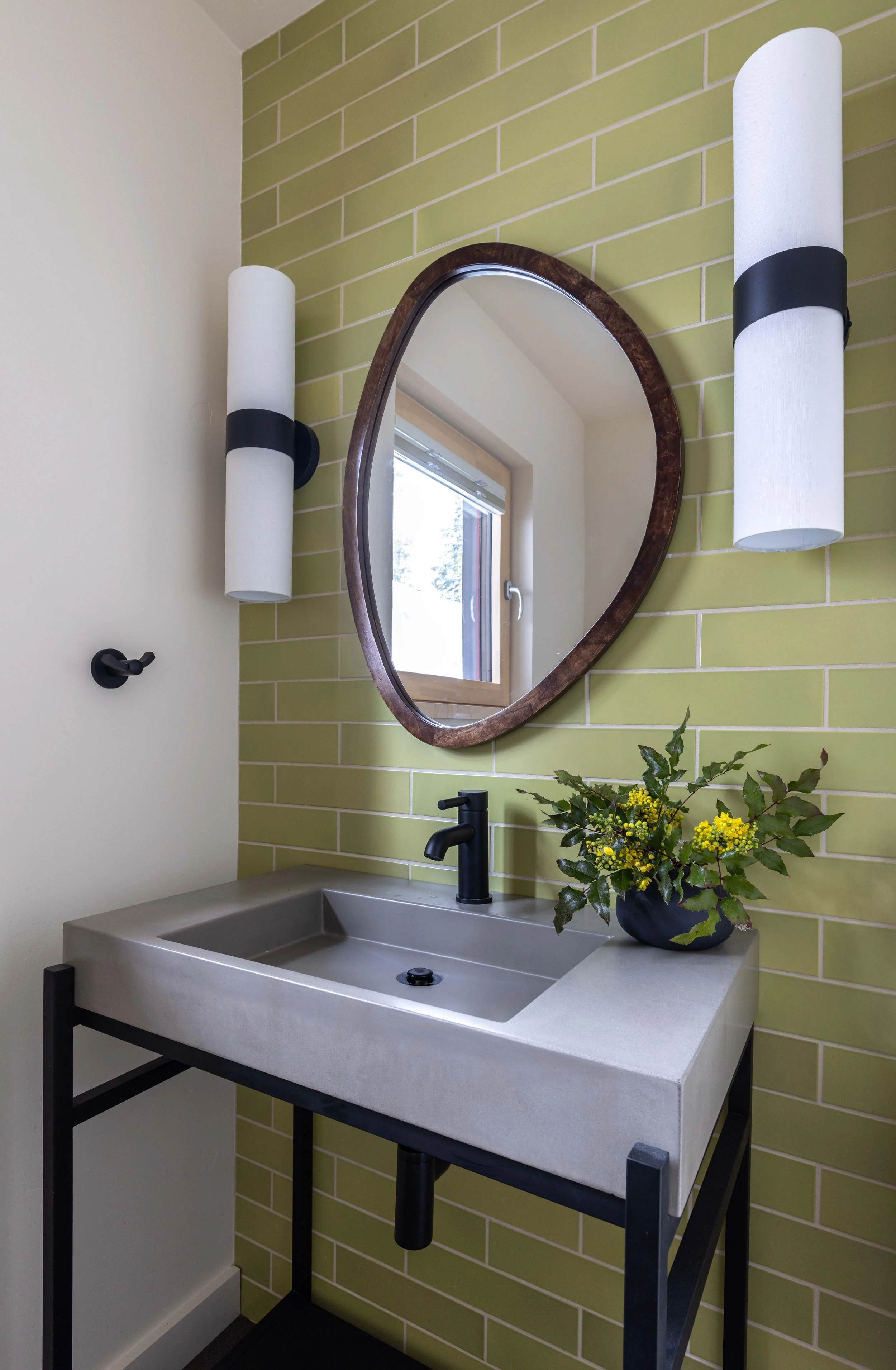 Modern bathroom with green tile wall, oval mirror, concrete sink, and wall sconces. Black faucet and vase with greenery on sink.