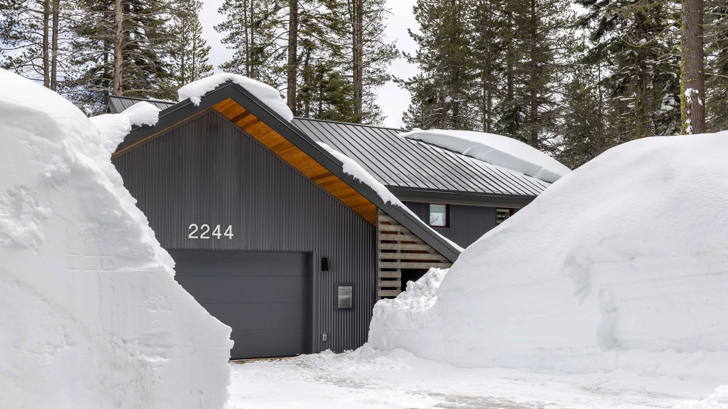 Modern house with metal roof surrounded by deep snow, trees in the background.