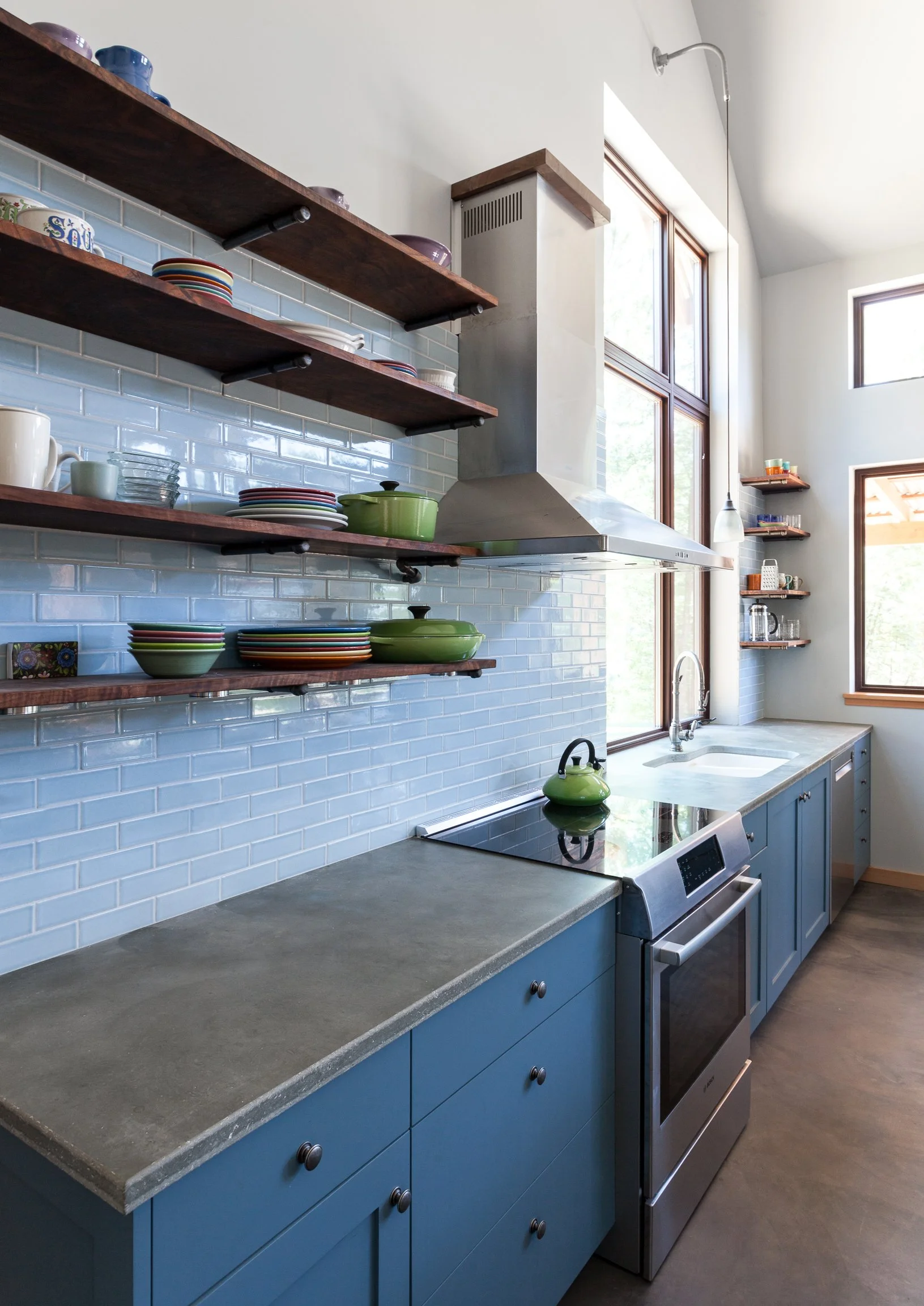 Modern kitchen with blue cabinetry, concrete countertops, and stainless steel appliances. Open wooden shelves hold colorful dishes and a green kettle sits on the stove. A large window brings in natural light.