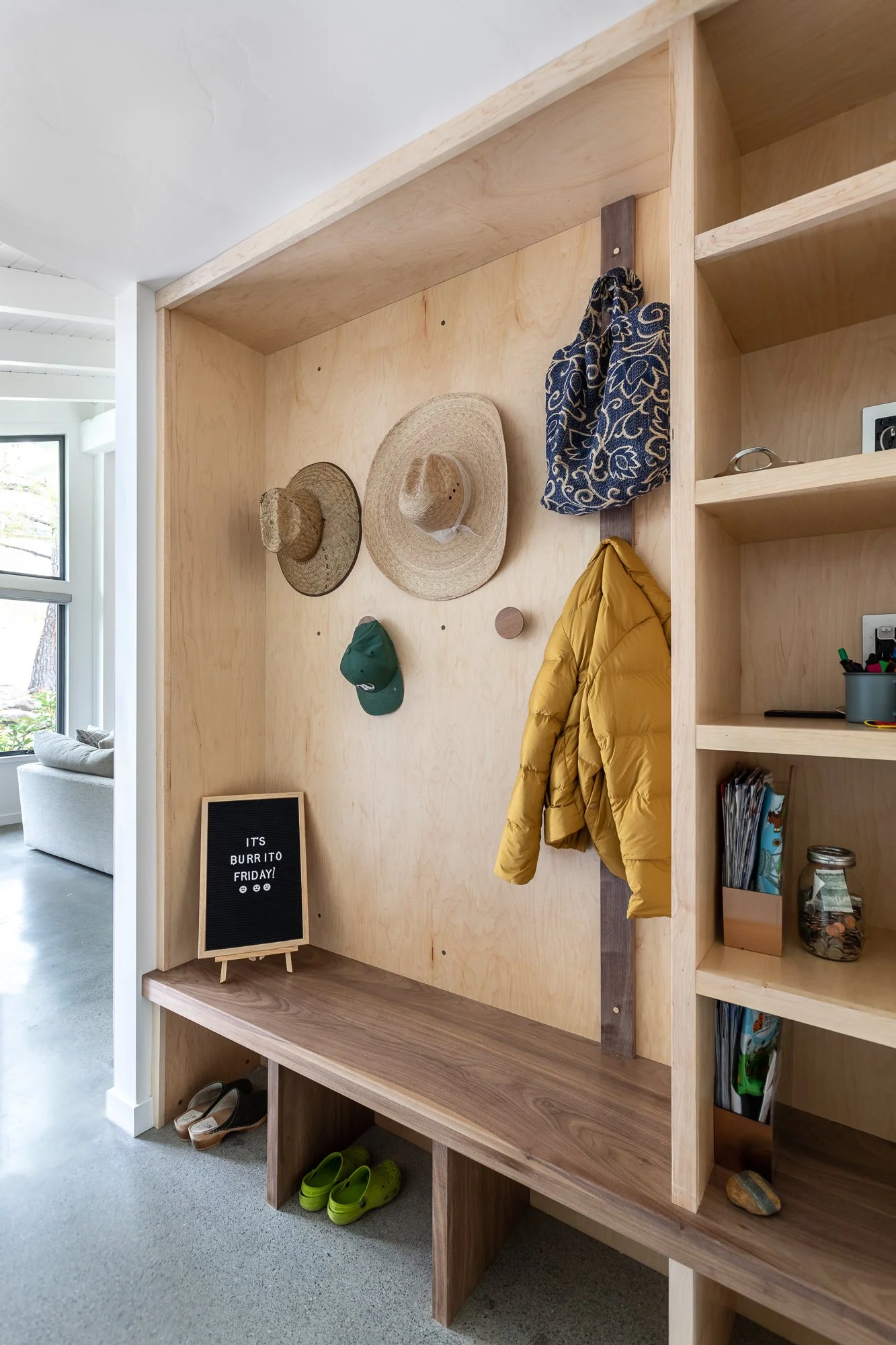 Entryway with wooden shelving, hooks holding straw hats, a green cap, a patterned bag, and a yellow jacket. A felt letter board reads 'It's Burrito Friday!' on a wooden bench. Shoes are placed below, and shelves contain various items like magazines and a jar.