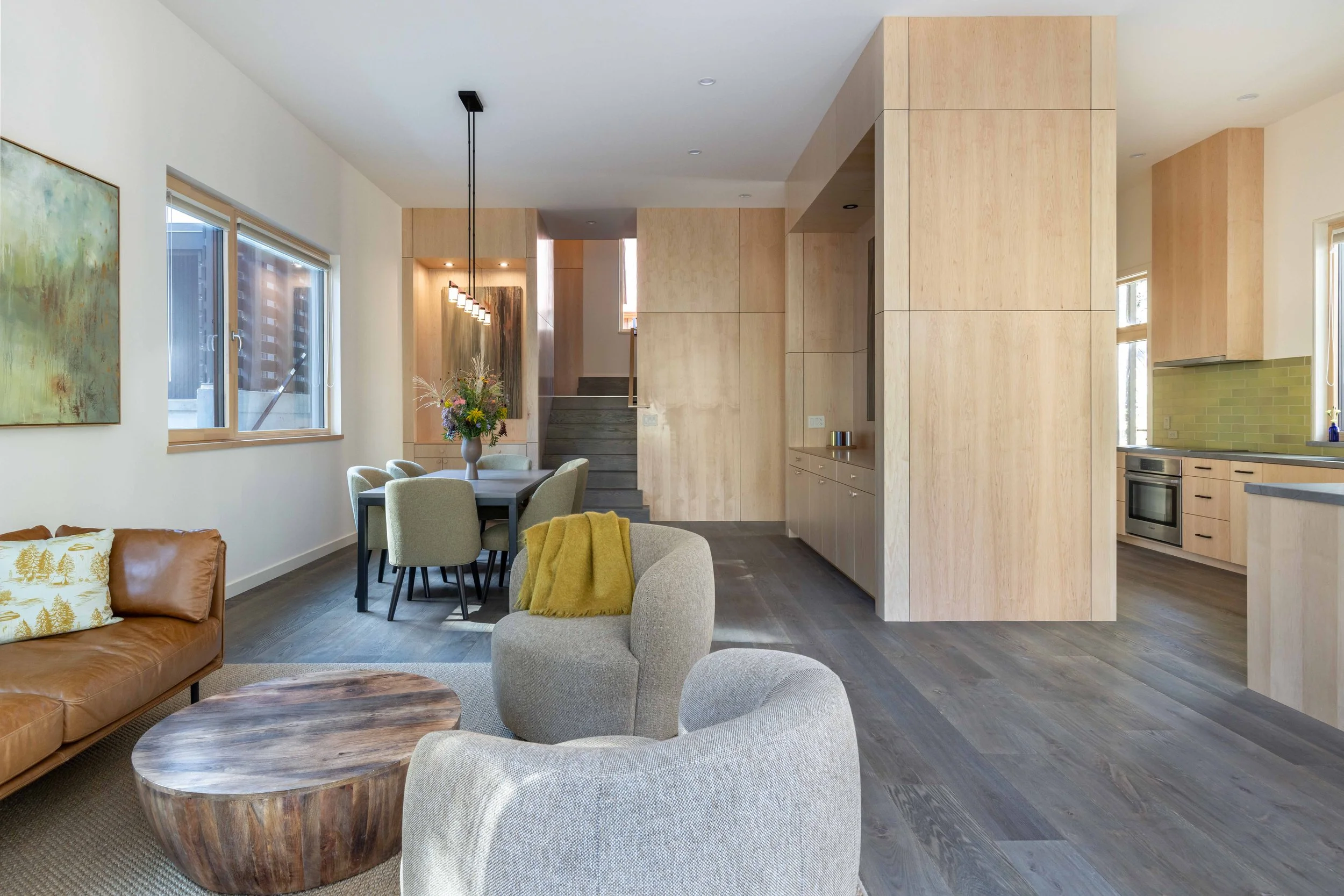Modern open-plan living and dining area with light wood decor, featuring a tan leather sofa, textured gray chairs, a round wooden coffee table, and a dining table with chairs. A minimalist kitchen area with light wood cabinets and a green tile backsplash is visible. Large windows provide natural light and there is modern artwork on the wall.