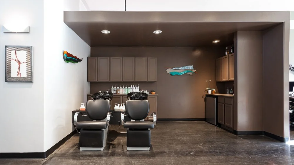 Interior of a modern hair salon with two shampoo chairs facing cabinetry and shelves holding hair care products.