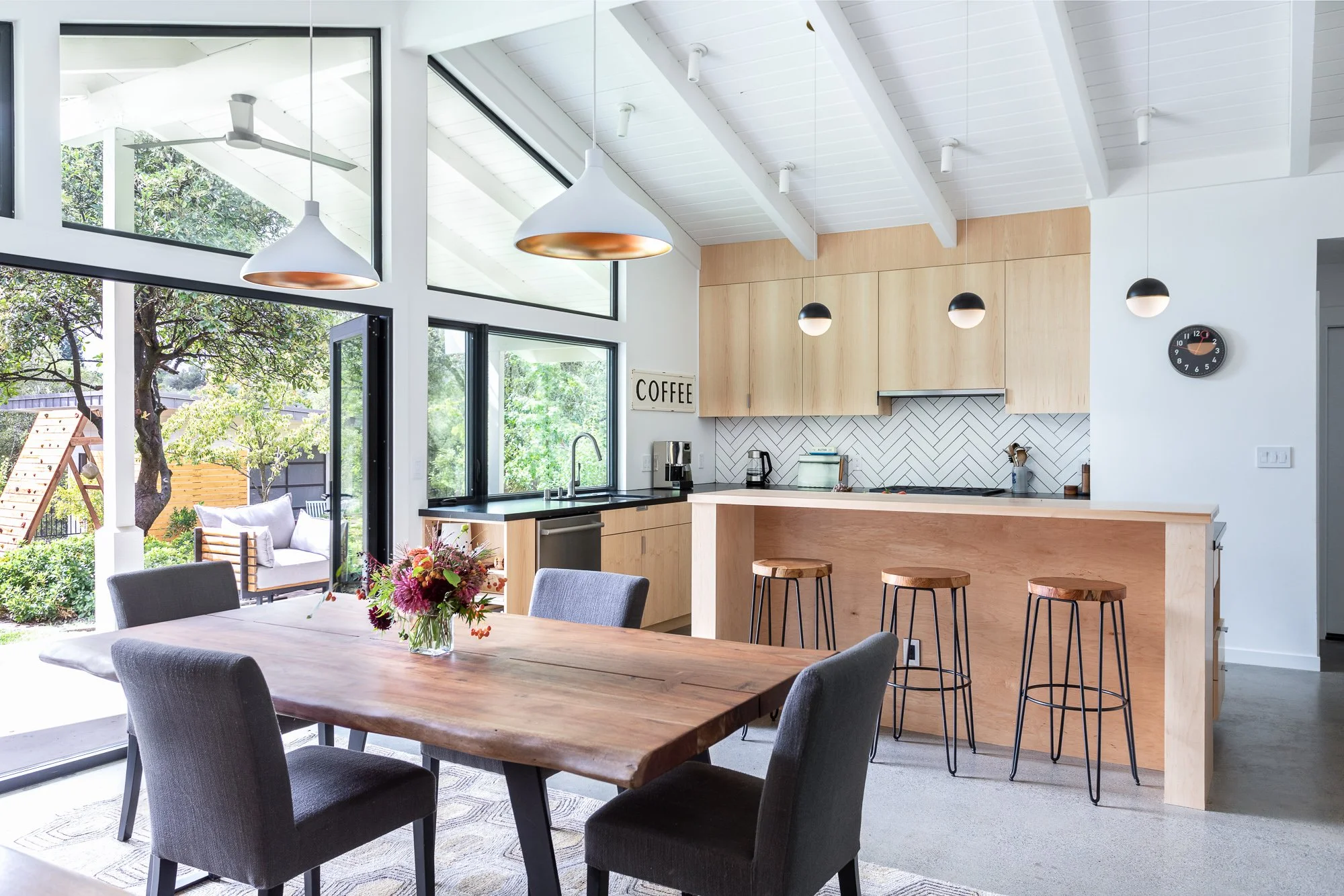 Modern open-concept kitchen and dining area with wooden table, gray chairs, minimalist wood cabinets, pendant lighting, bar stools, and large windows overlooking a garden.