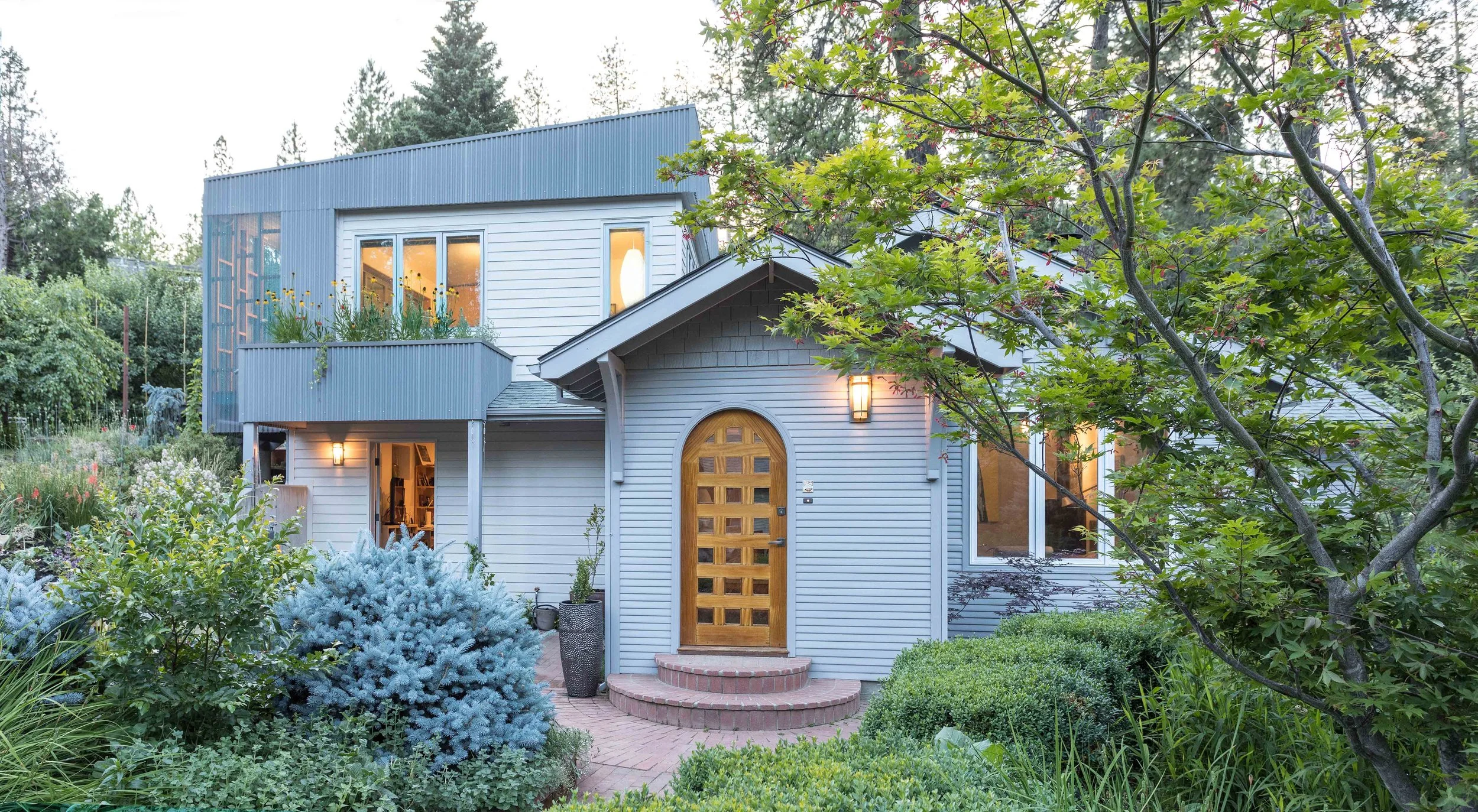 Modern two-story house with lush garden, featuring a wooden arched front door, large windows, and a balcony with plants.