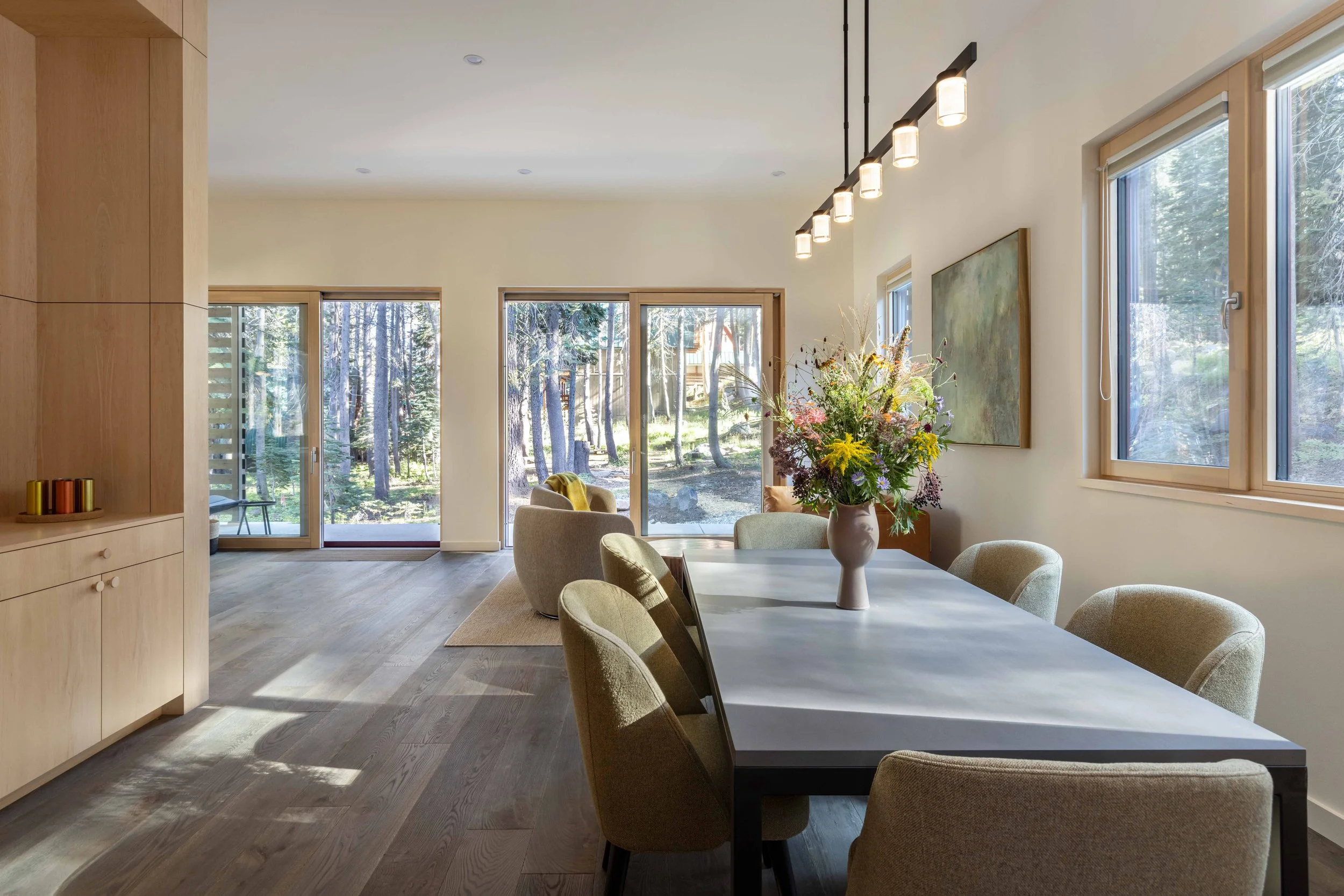 Modern dining room with a large table, upholstered chairs, and a floral centerpiece. Large windows and a sliding door provide views of a wooded outdoor area. Light wood cabinetry and flooring complement the bright, open space.