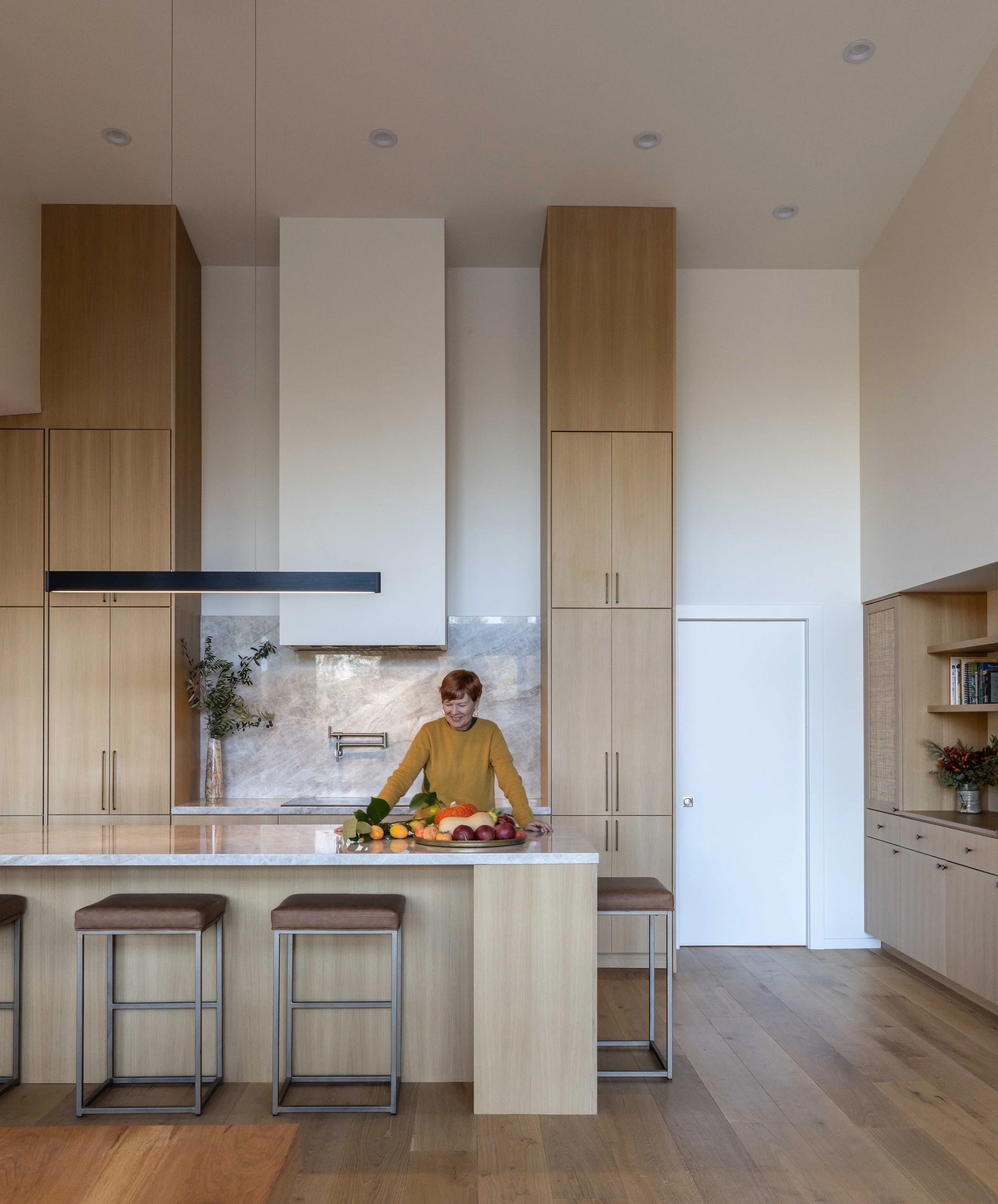 Modern kitchen with high ceilings, wooden cabinets, marble countertop, bar stools, and a person organizing fruits on the island.