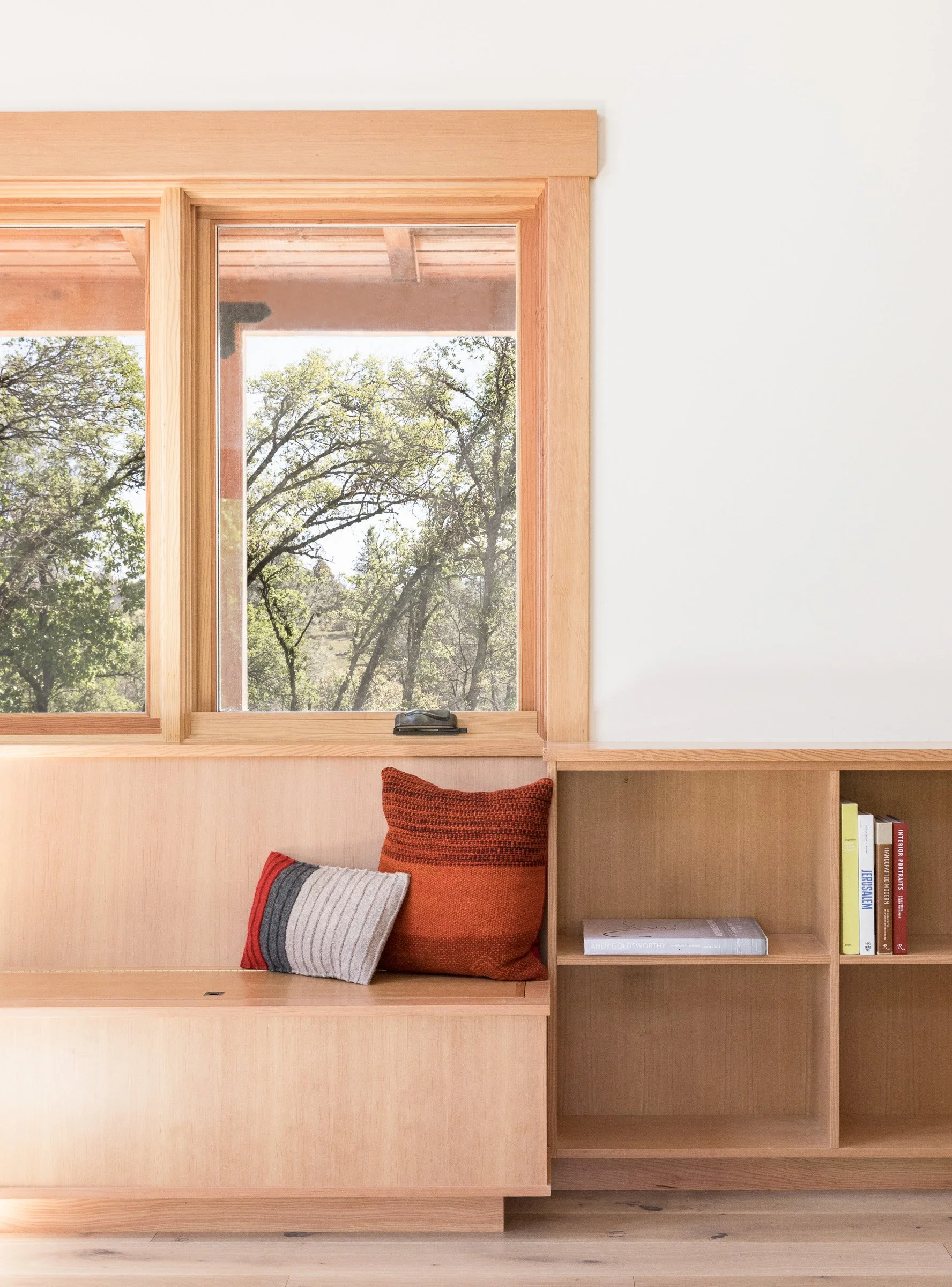 Wooden window seat with red and gray cushions next to a bookshelf, with a view of trees through the window.