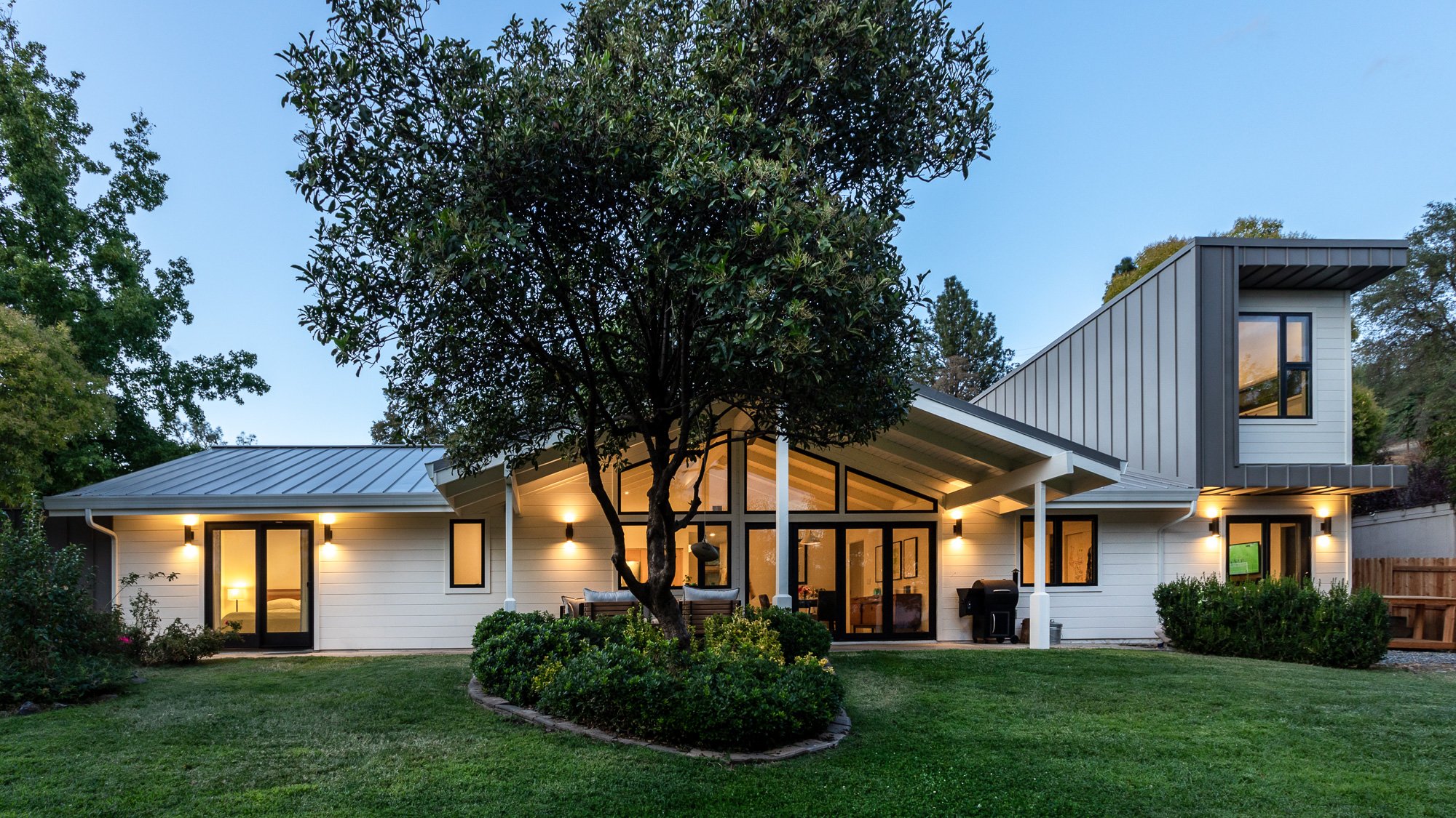 Modern house exterior with large windows, sloped metal roof, and surrounding trees at twilight. Lawn and garden in the foreground.
