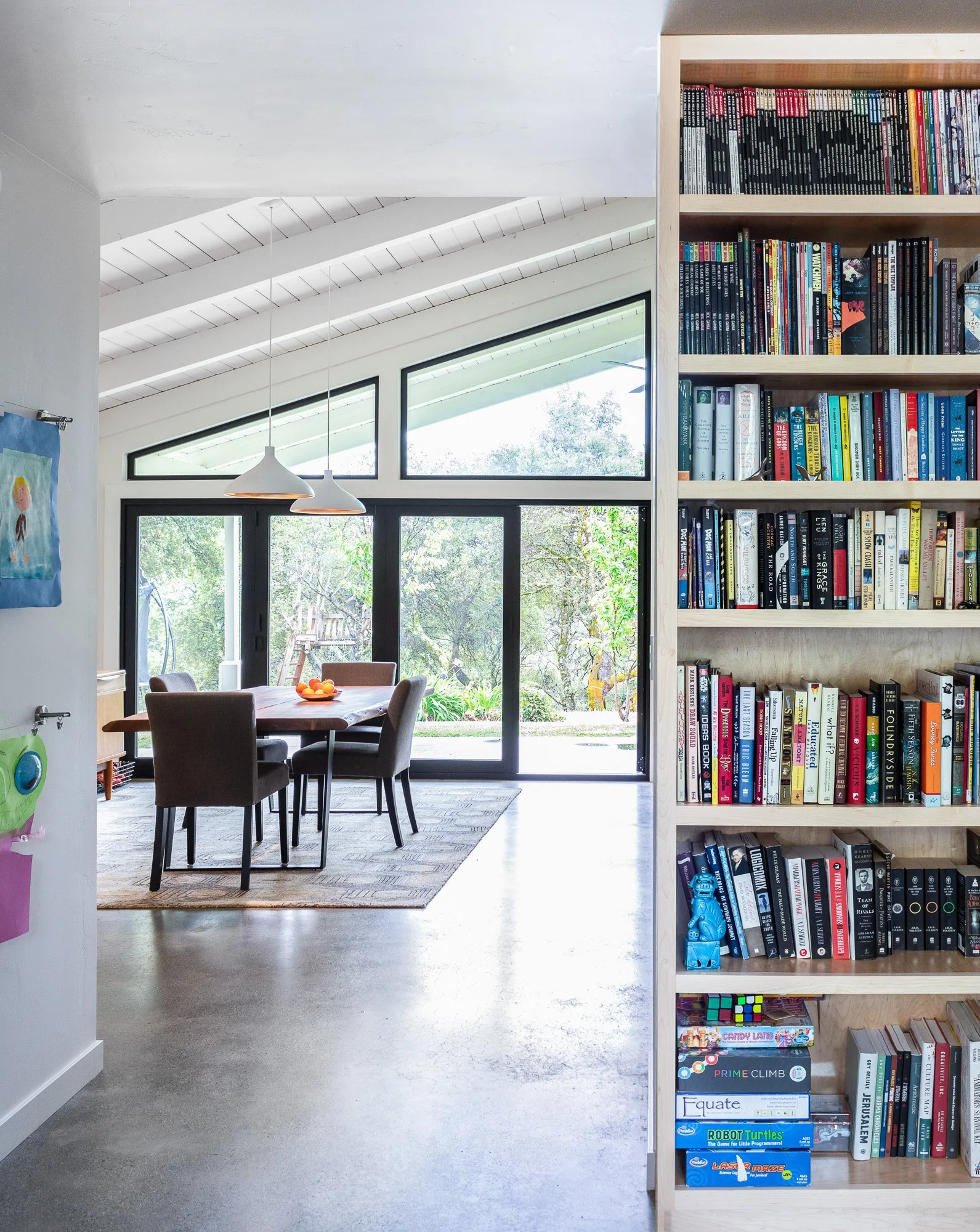 Modern living space with large windows, dining table with chairs on a rug, bookshelf filled with books and games, pendant lights, and a view of trees outside.