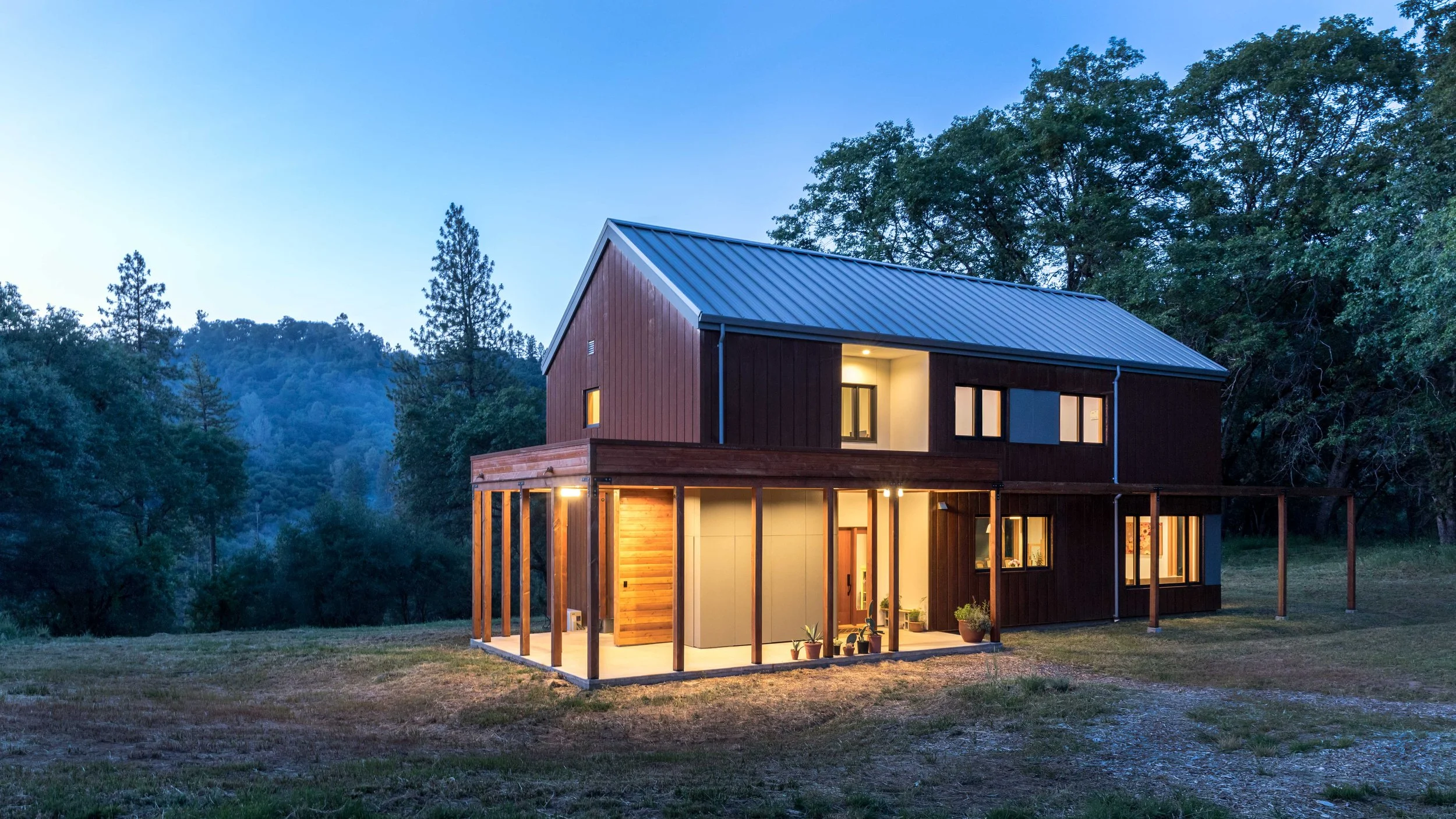 Modern wooden house with metal roof, surrounded by trees and grassland, during twilight.